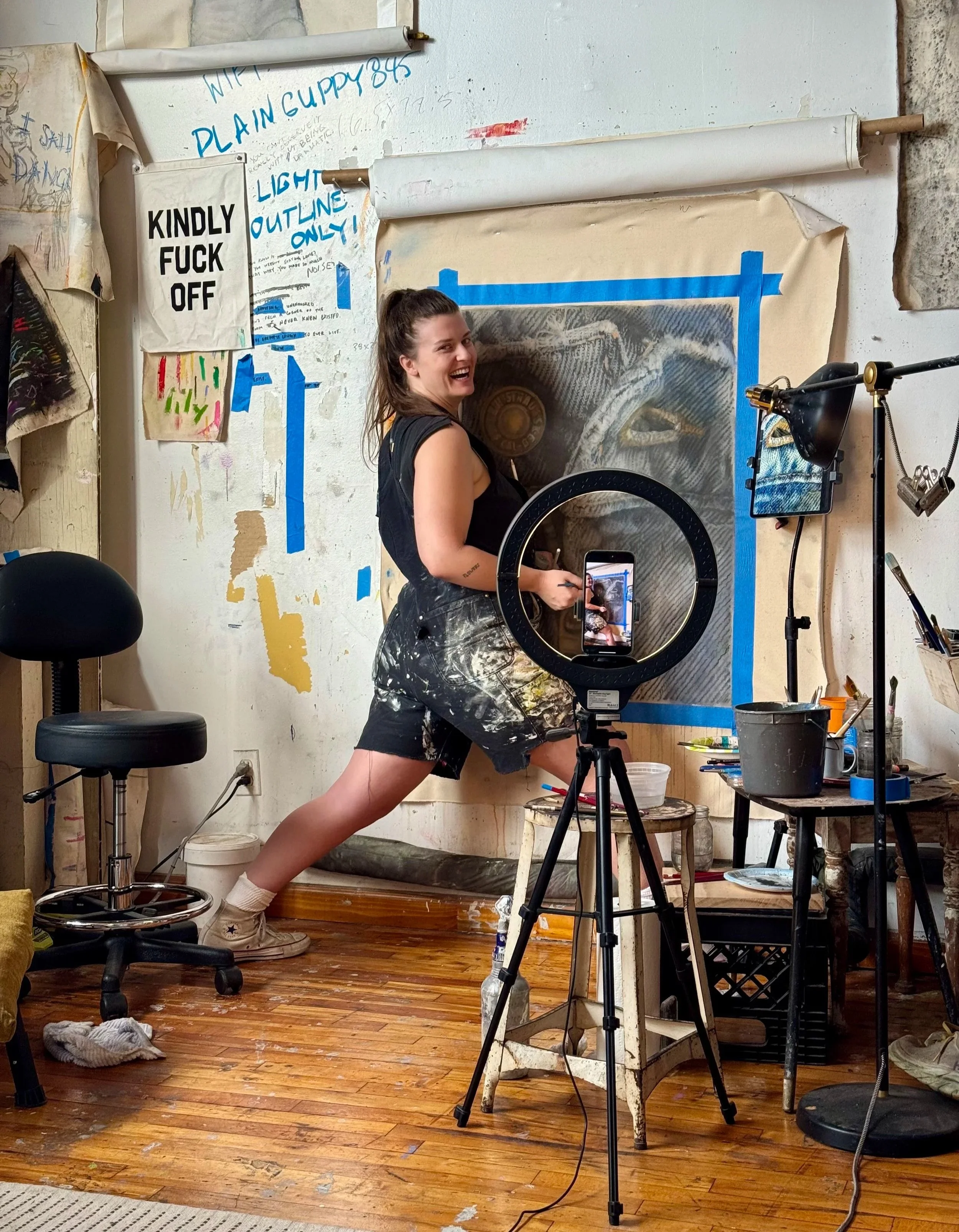 An art studio with a young woman smiling in front of an artwork, surrounded by art supplies and equipment.