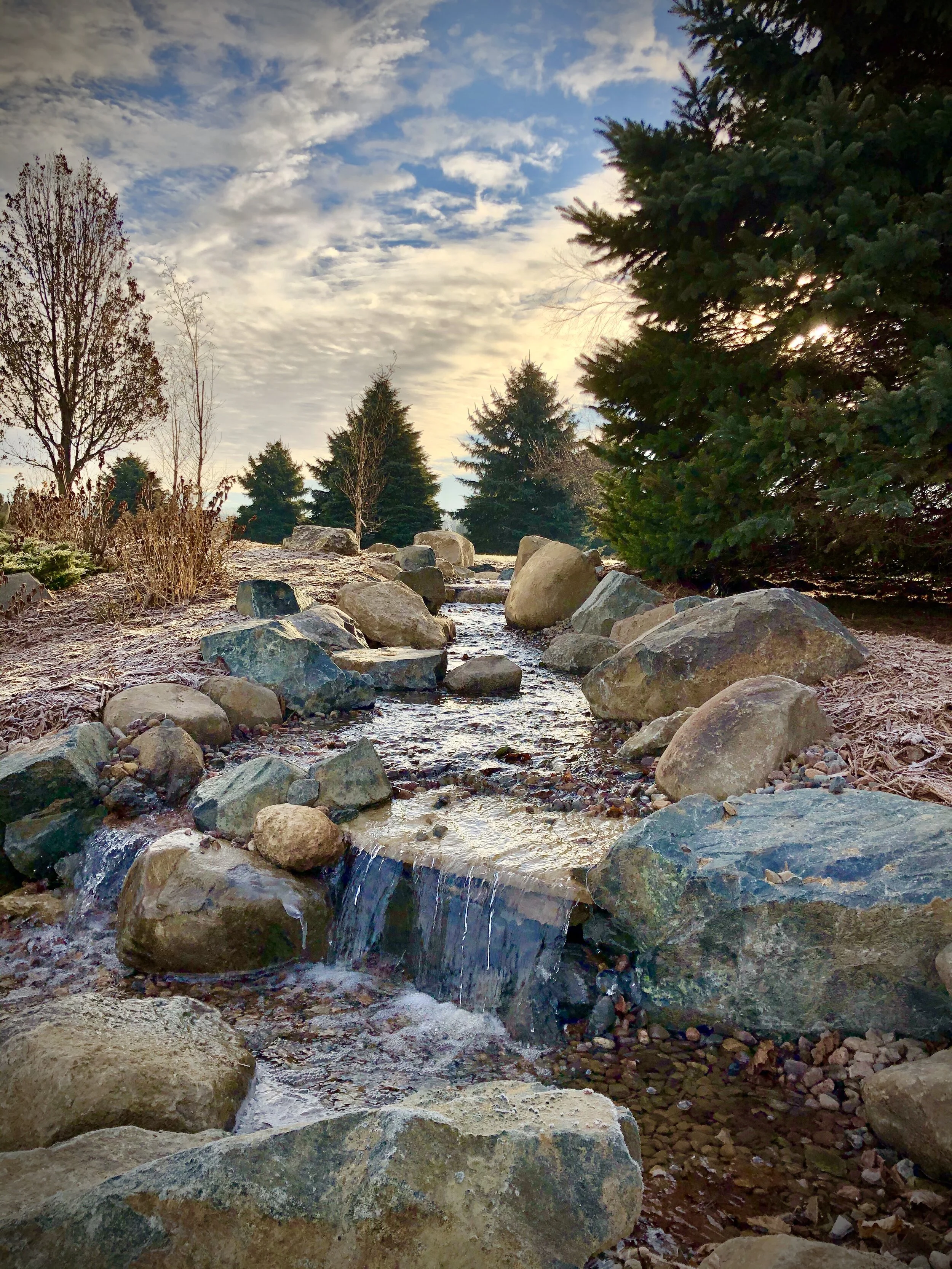A small stream flowing over rocks in a natural outdoor setting with trees and a cloudy sky at sunrise or sunset.