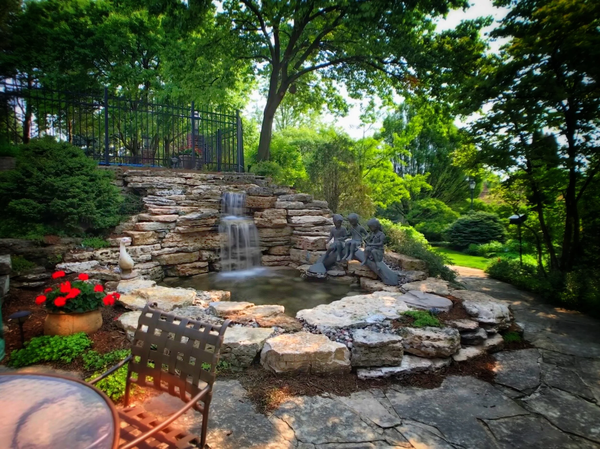 A landscaped backyard garden with a small waterfall cascading over stacked rocks, a sculpture of three children sitting together, and lush green trees and bushes. There is a black metal fence at the top of the rocks, and a bird statue near red flower
