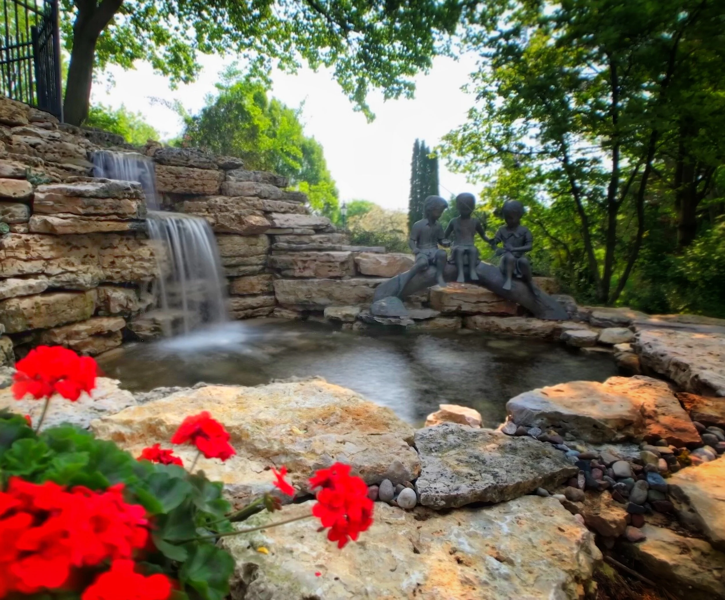 A garden scene with a small waterfall cascading over a stone wall, a bronze sculpture of three children sitting together, surrounded by lush green trees and vibrant red flowers in the foreground.