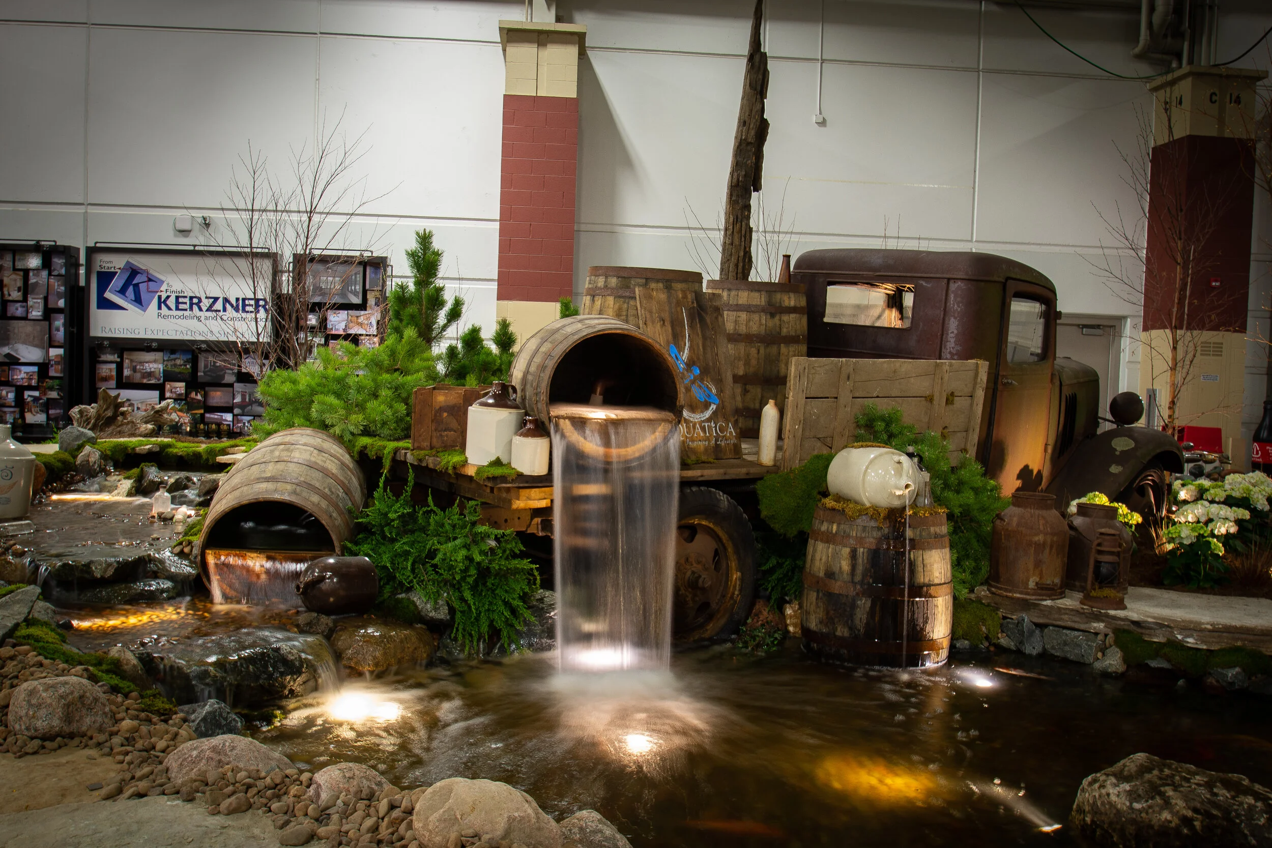 Indoor display featuring a miniature landscape with a small water stream flowing from a rustic truck-shaped water feature, surrounded by rocks, small trees, and decorative objects, with informational posters in the background.