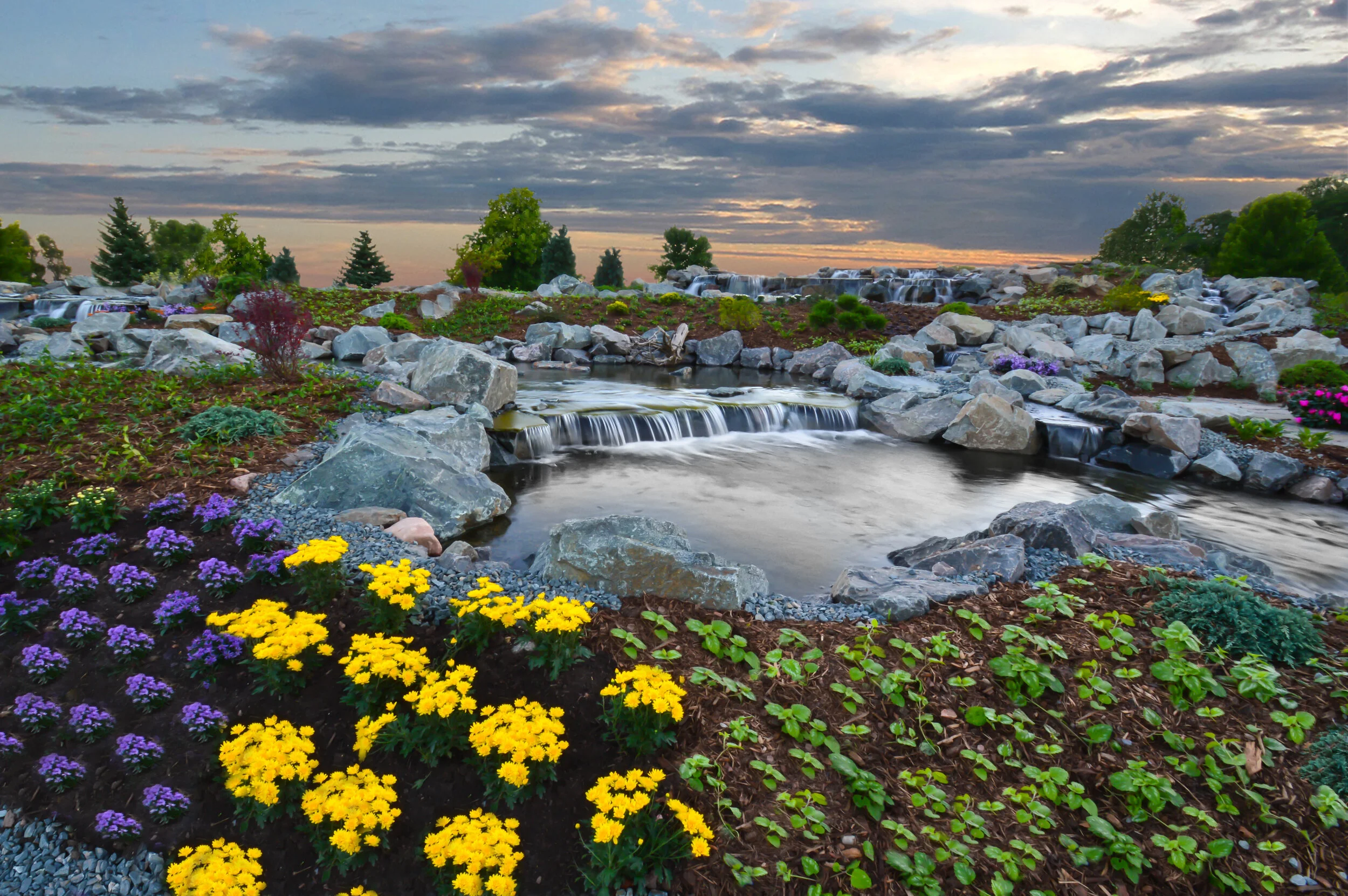 A landscaped garden with colorful flowers, rocks, a small waterfall, and a pond under a cloudy sky at sunset.