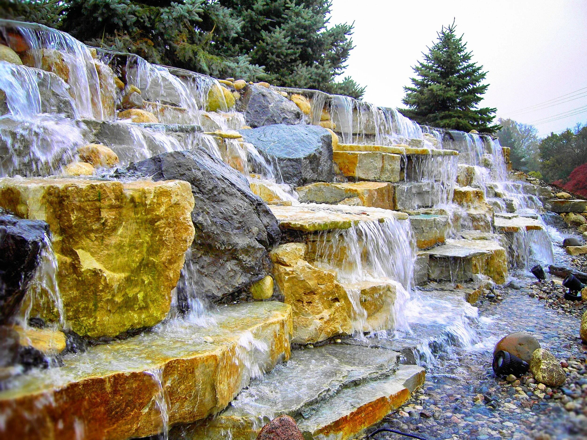Water flows over a multi-tiered stone waterfall surrounded by trees, rocks, and pebbles in a natural outdoor setting.