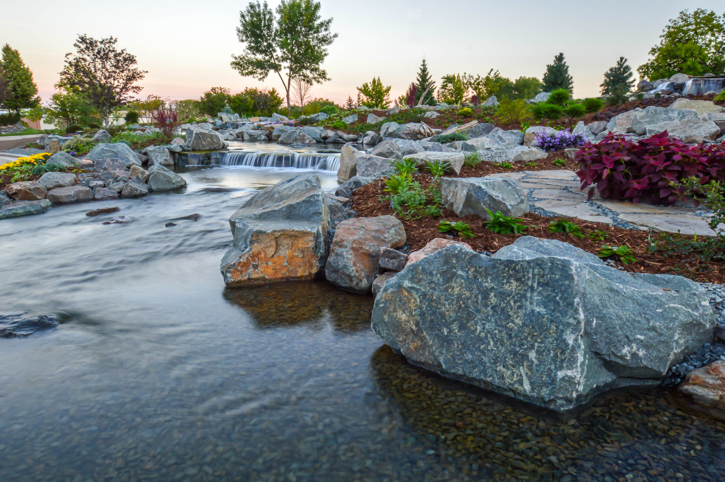 A landscaped garden with a small waterfall flowing over rocks, surrounded by colorful flowers and lush green trees during sunset.