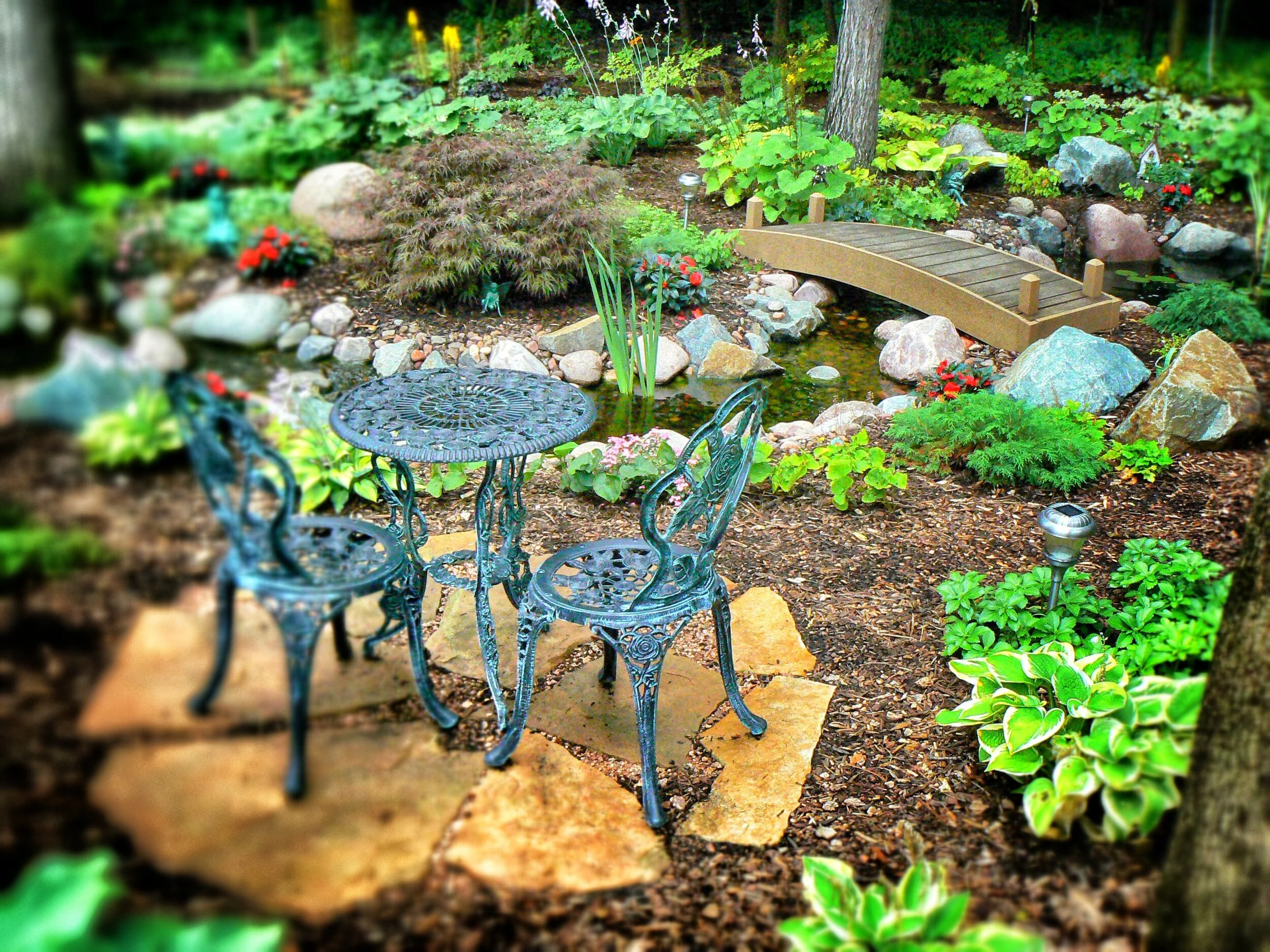 A garden with a small arched wooden bridge over a pond, surrounded by lush green plants, rocks, and decorative garden lights.