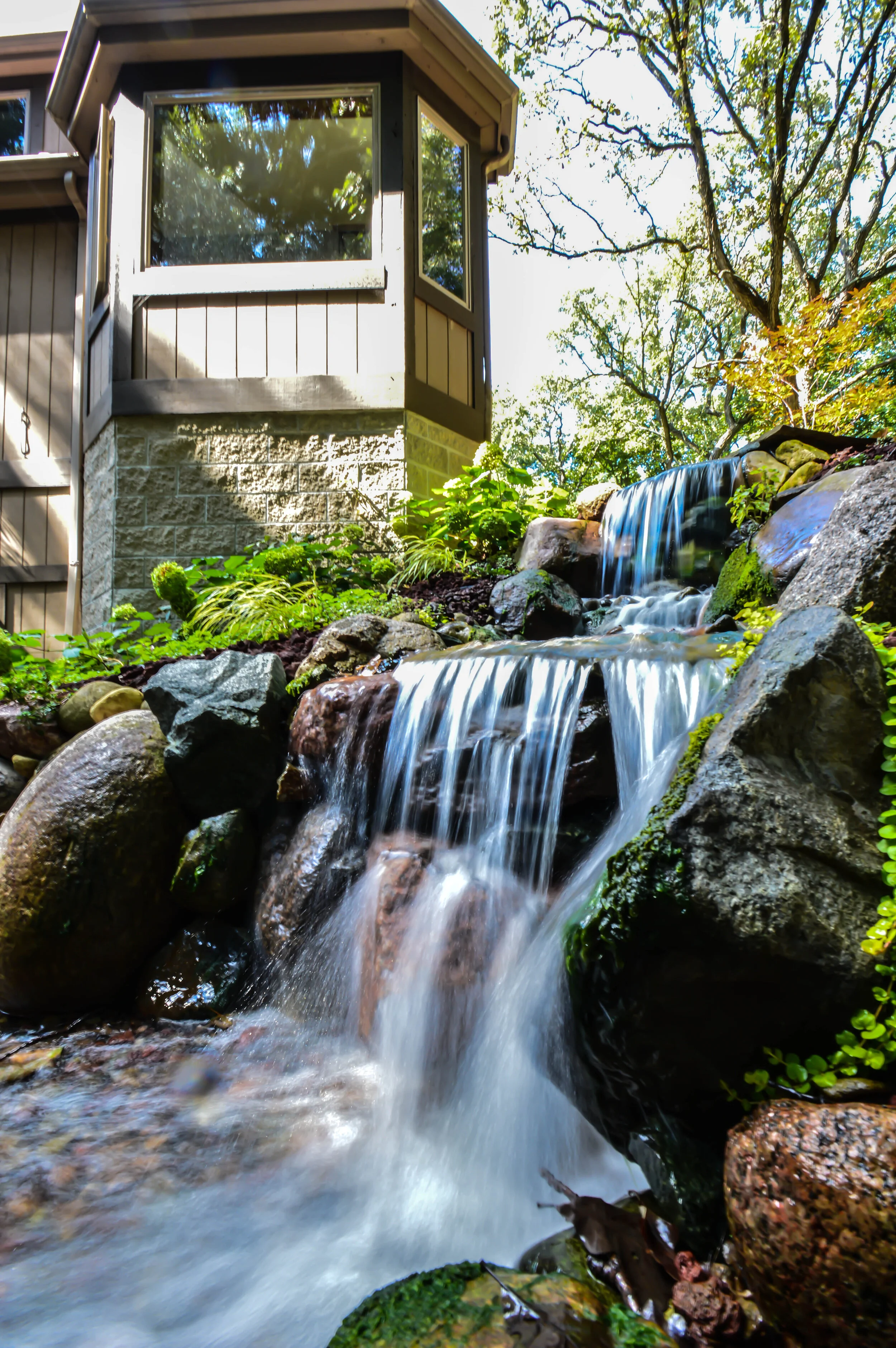 Small waterfall flowing over rocks beside a house with large windows, surrounded by trees and greenery.