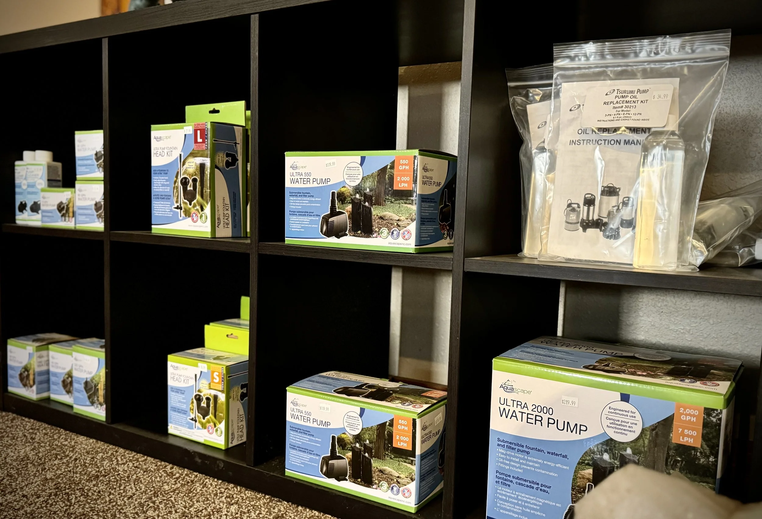 Black shelving unit displaying water pumps and related equipment, with boxes labeled 'Ultra 550 Water Pump' and 'Ultra 2000 Water Pump' and instruction manuals inside some packages.