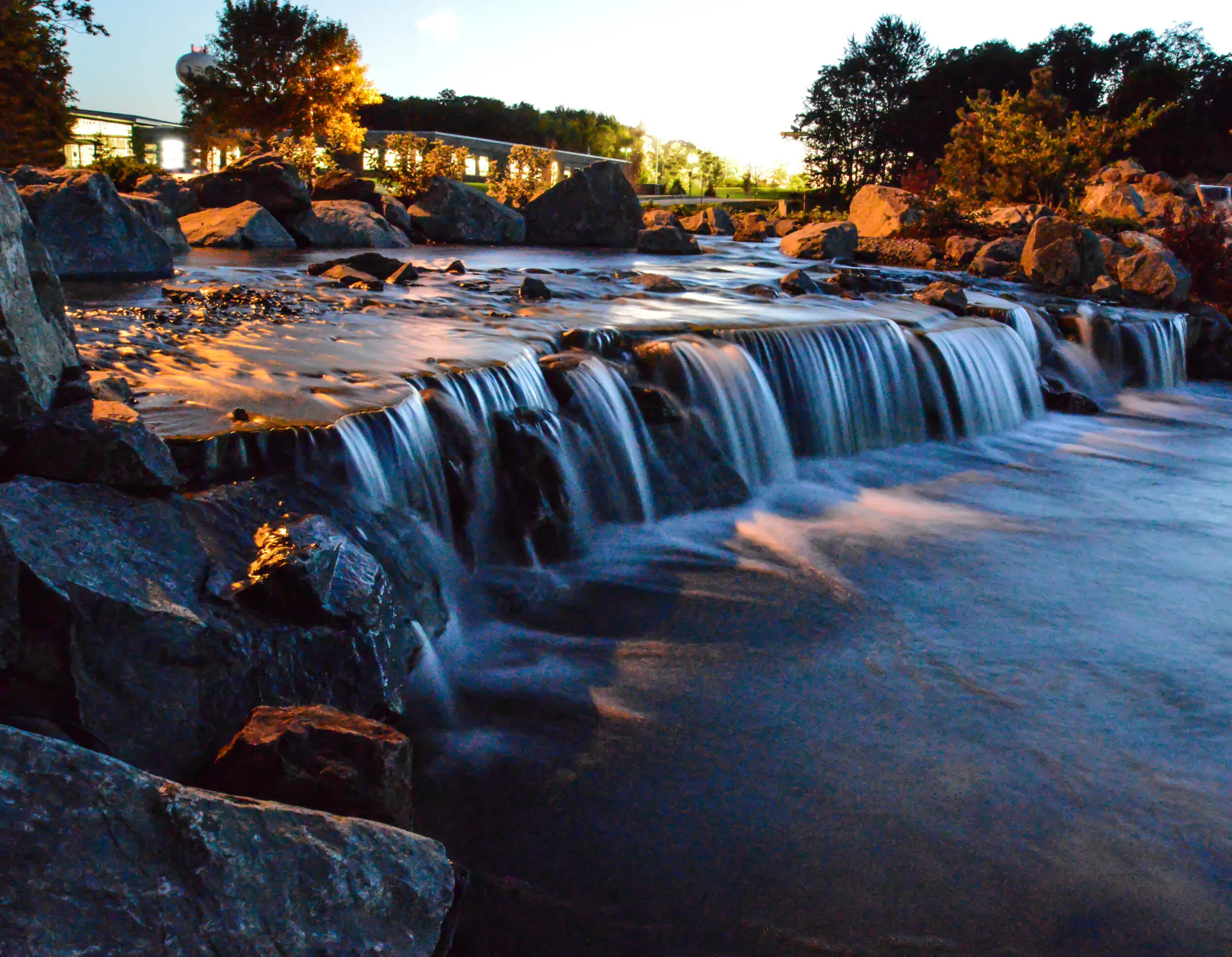 A small waterfall over rocks with a river flowing underneath, trees and buildings in the background, sky in twilight, and lights illuminating the scene.