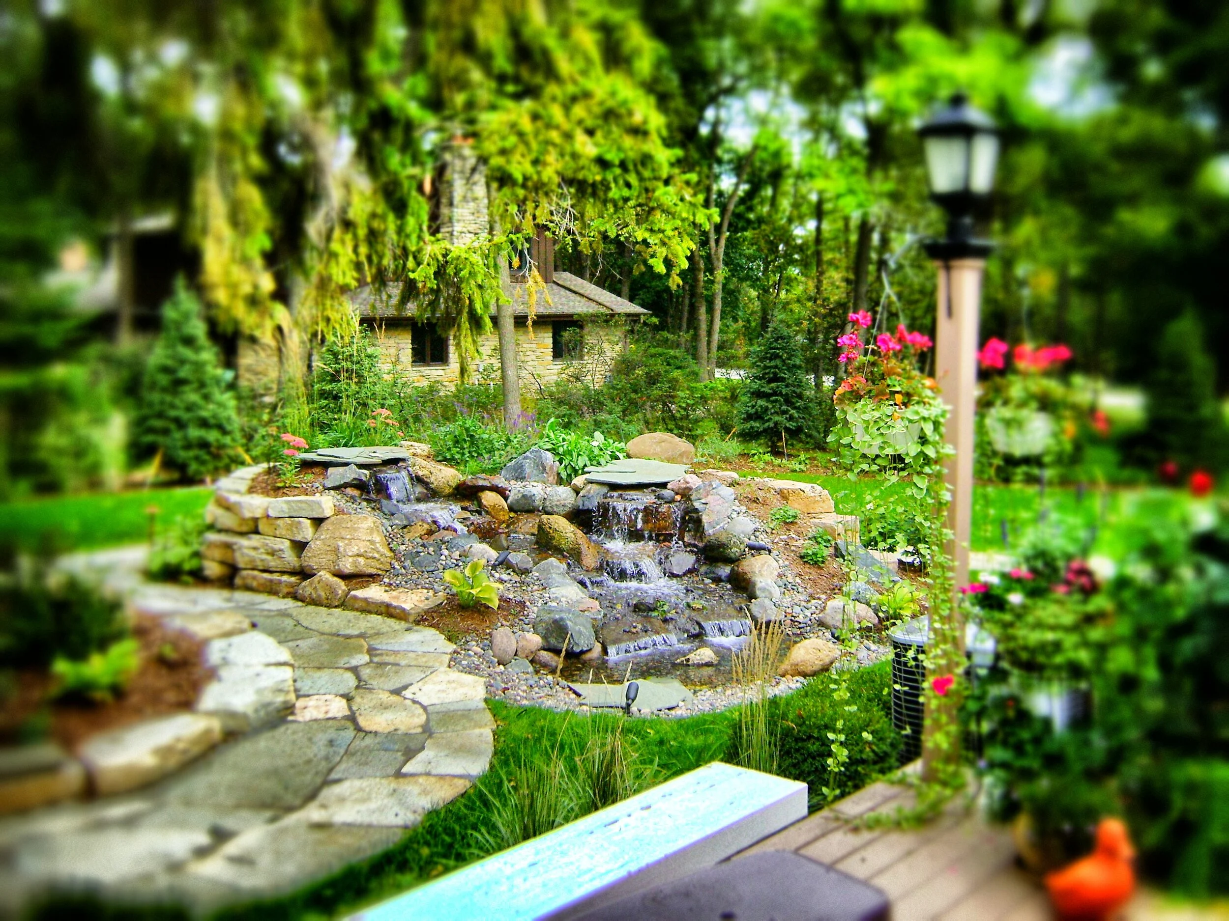 A lush green garden with a small stone waterfall feature, surrounded by various flowering plants and trees. There is a stone cottage with a shingled roof partially visible in the background, and garden lamps along the pathway.