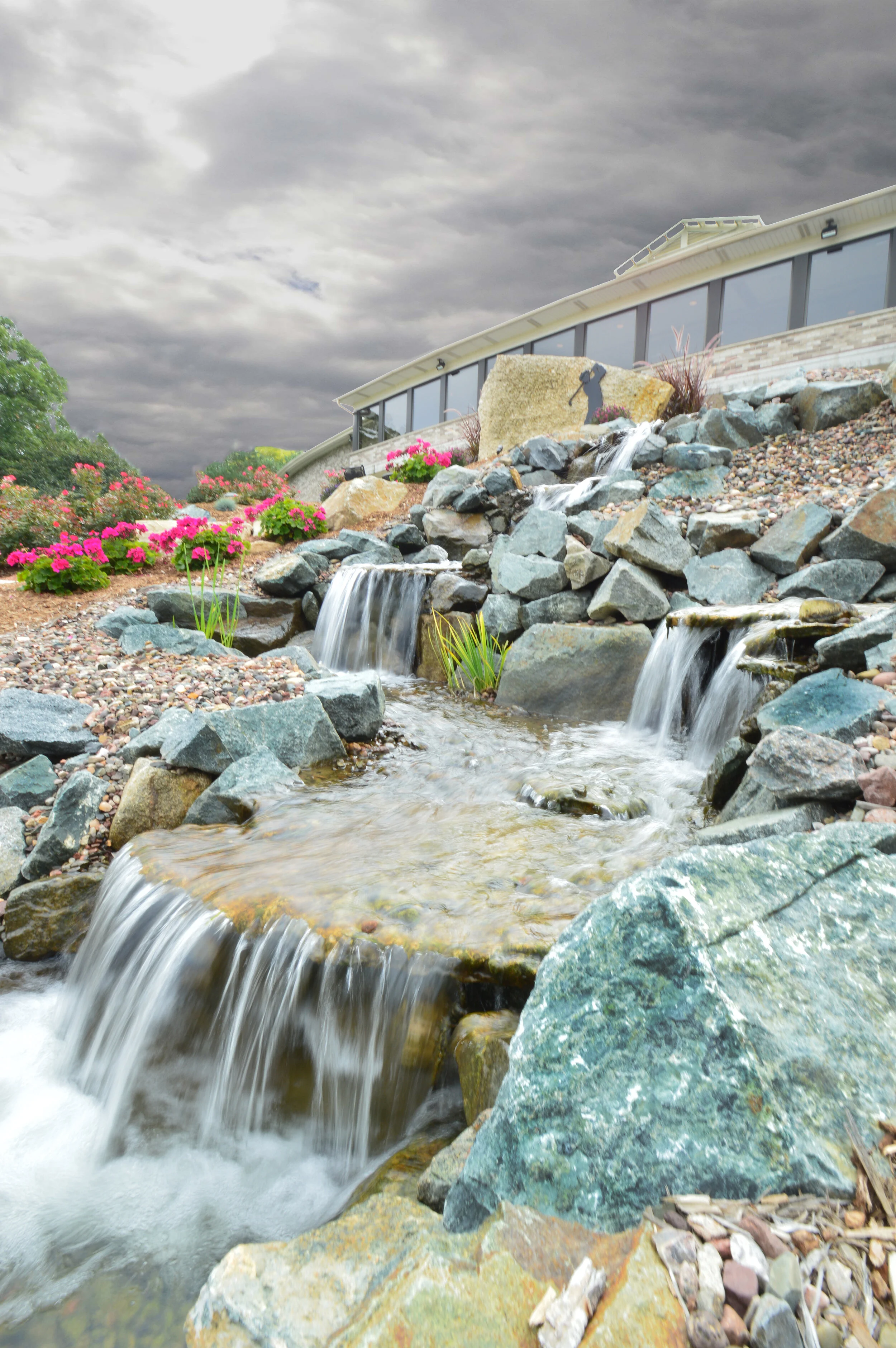 A small cascading waterfall flowing over rocks with a modern house with large windows in the background under a cloudy sky.