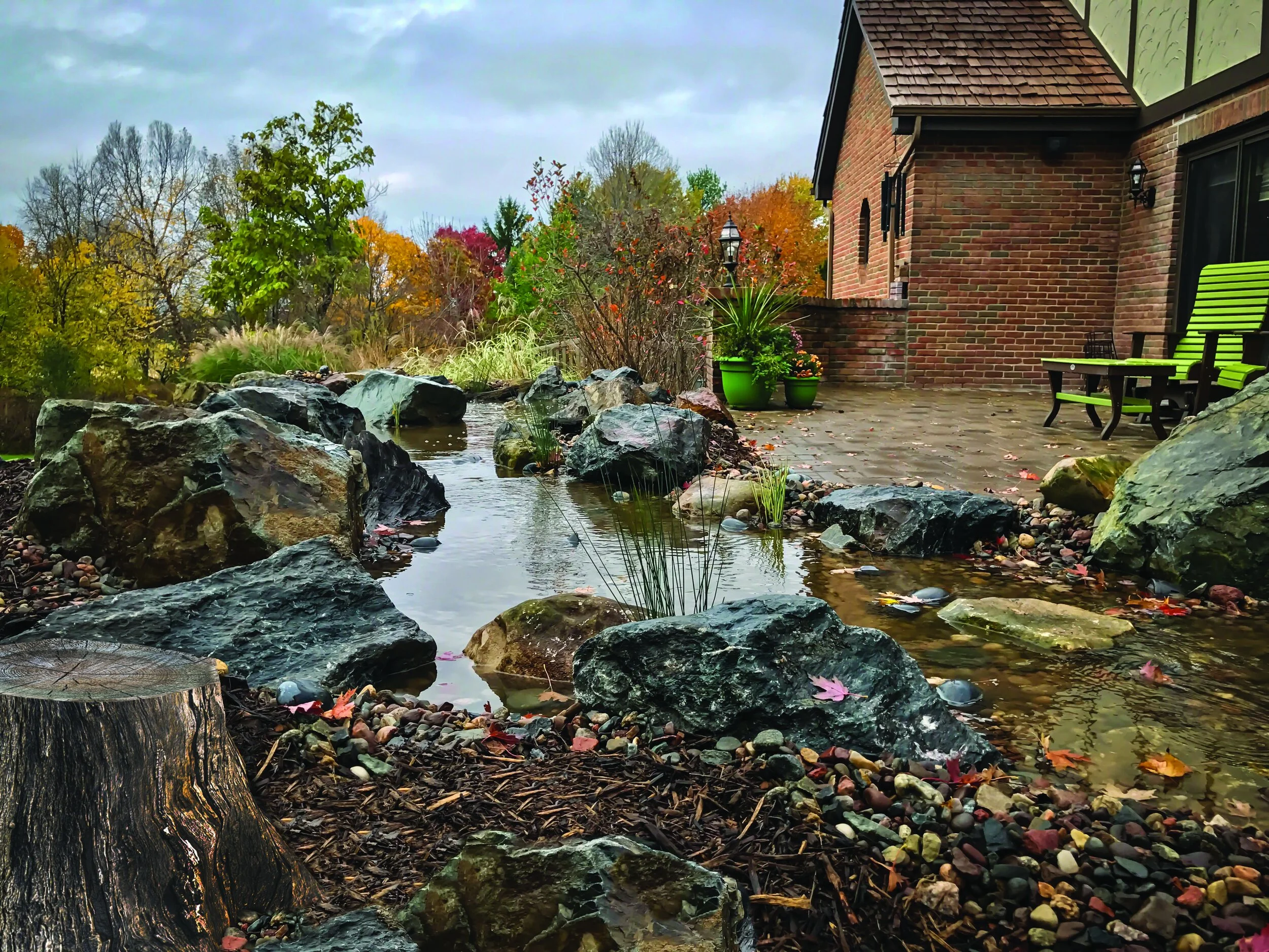 A backyard scene in autumn showing a brick house with a patio, green chairs, a garden with trees, shrubs, and a small stream with rocks and fallen leaves.