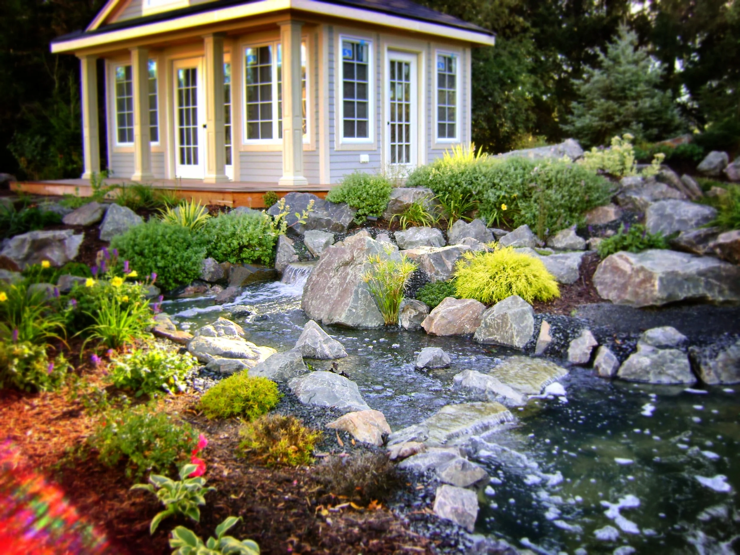 A small house with large windows surrounded by lush greenery and a garden with rocks and plants next to a flowing stream.