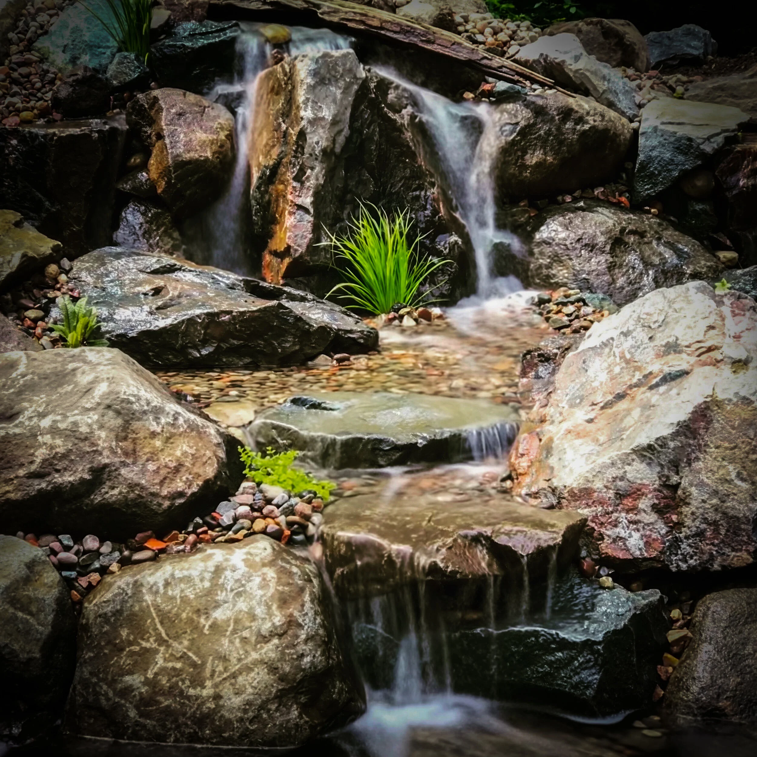 Small waterfall flowing over rocks in a natural setting, with green plants and pebbles around.