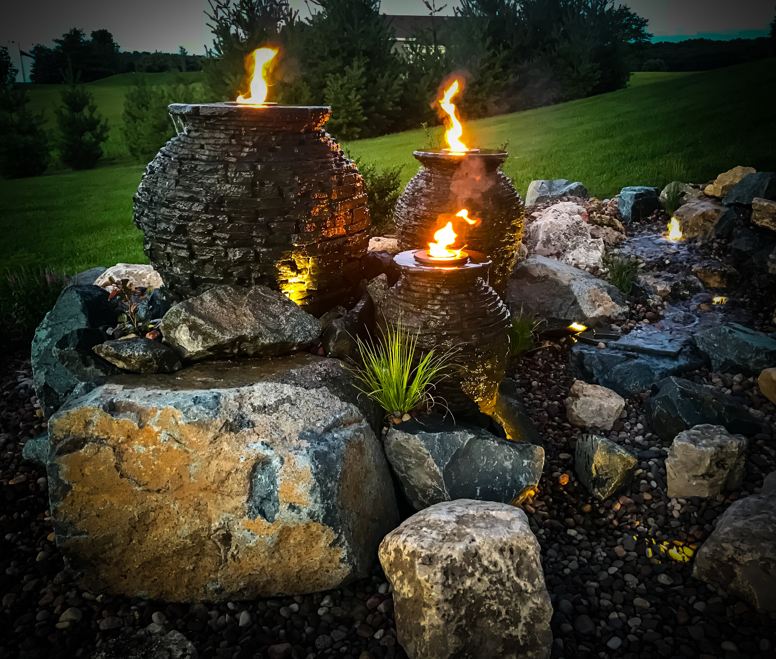 Three decorative fire bowls with flames are placed on a rocky landscape in a backyard at dusk. The background features green grass, trees, and a darkening sky.