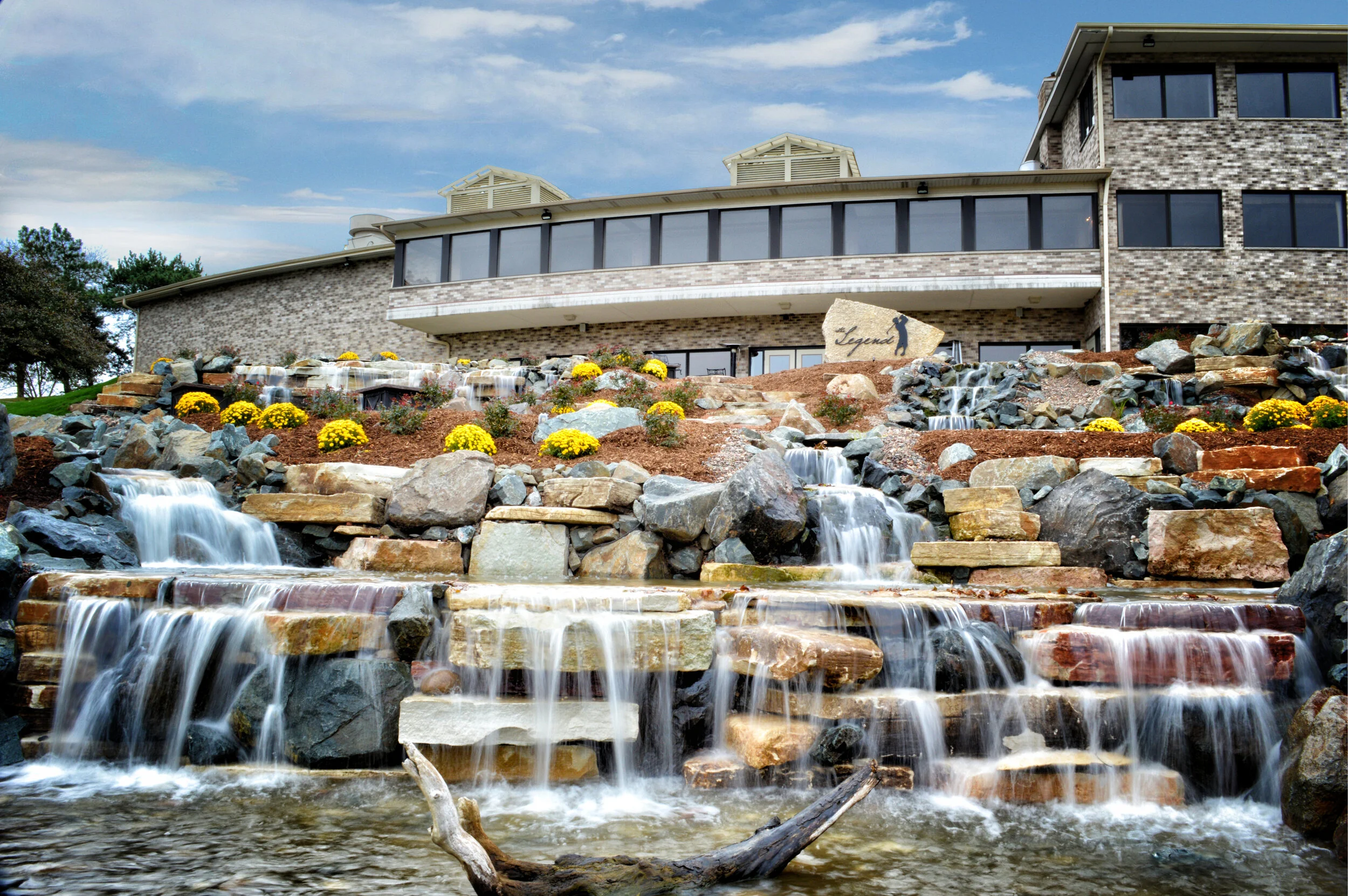 A modern house with a stone facade, large windows, and a landscaped garden featuring a waterfall with cascading rocks and yellow flowers.