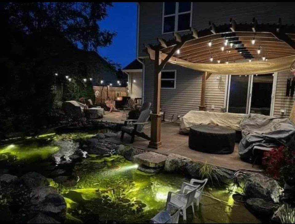 Backyard patio at night with a wooden pergola, outdoor seating, a small pond with glowing lights, and string lights overhead.