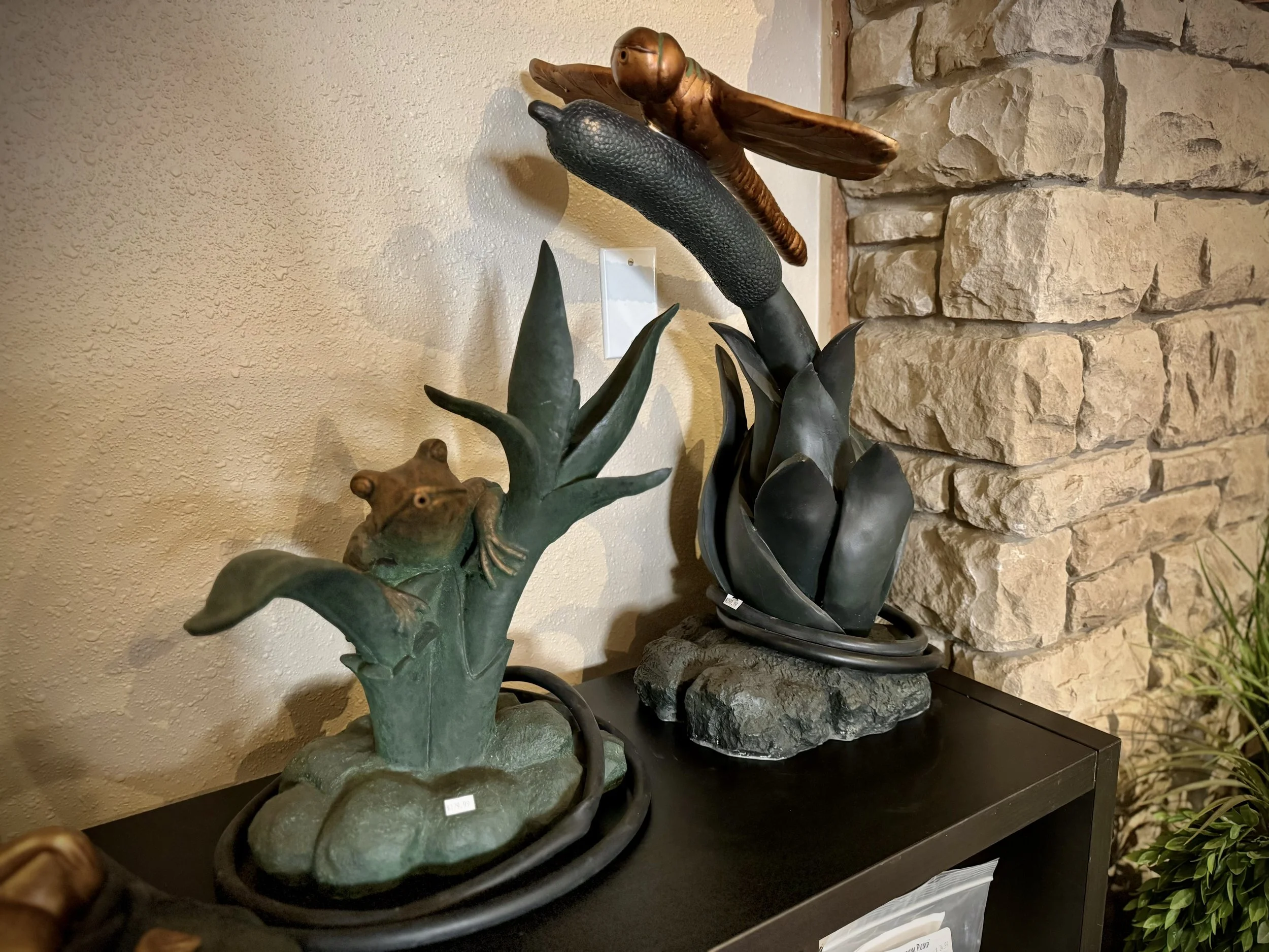 Decorative sculptures of frogs and plant leaves displayed on a black table against a beige wall and brick wall background
