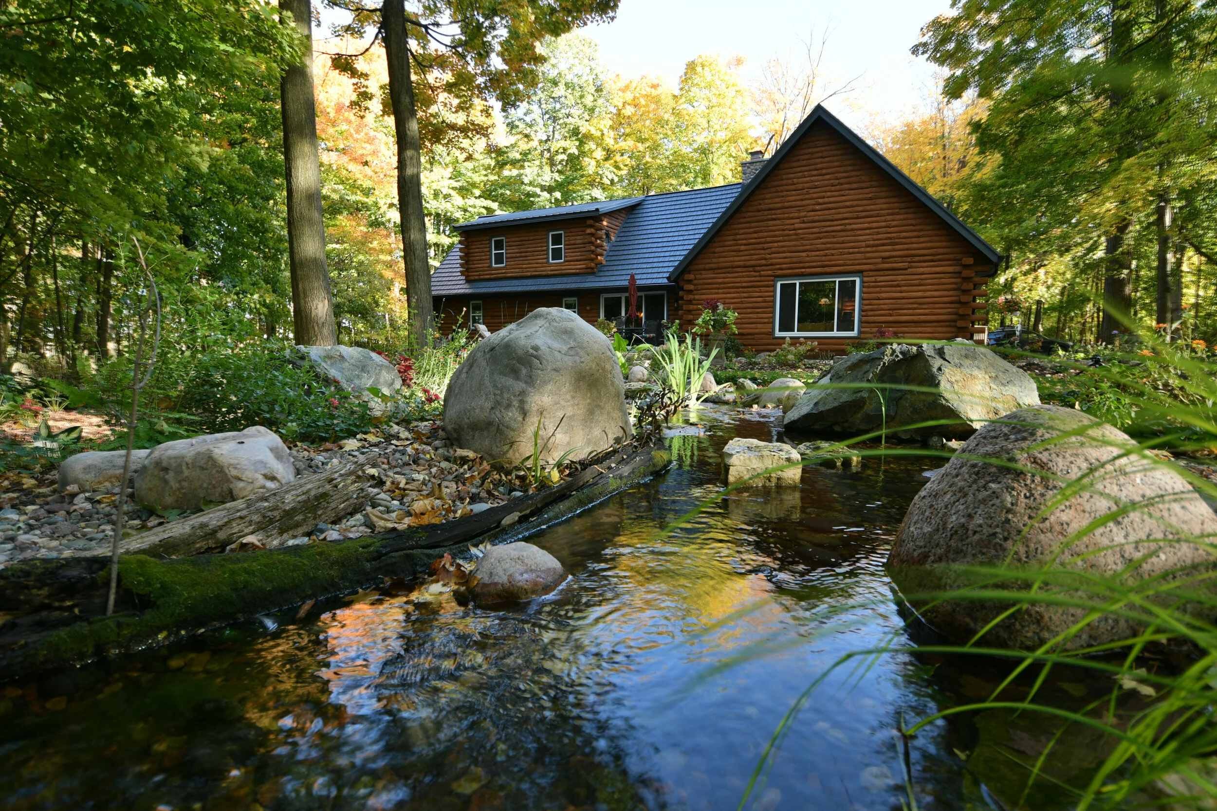 A rustic log cabin with a blue metal roof situated among lush green trees in a forest, with a small stream flowing in the foreground over rocks and pebbles.