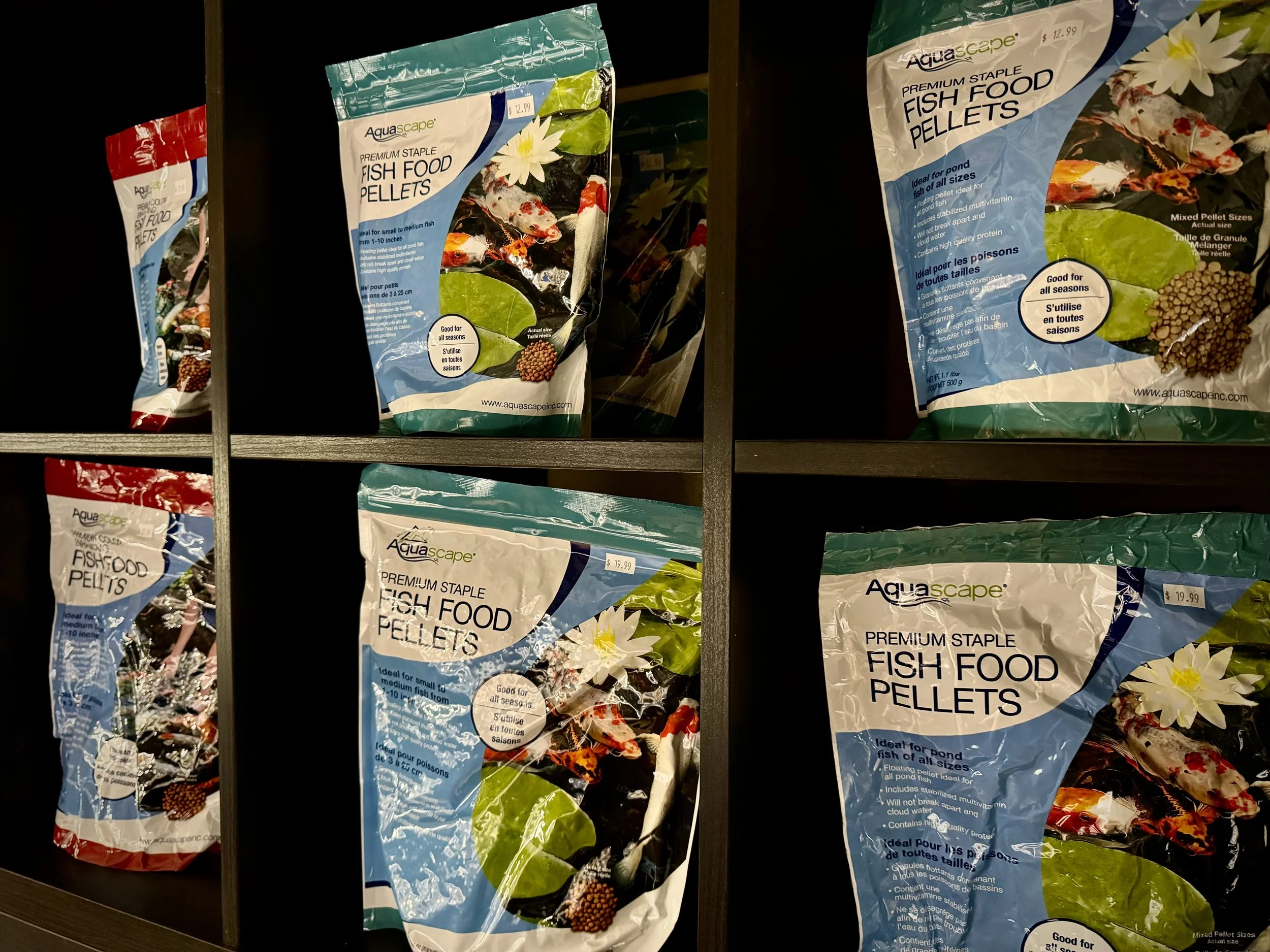 Multiple packages of Aquascape brand fish food pellets on a dark shelf. The packages are primarily blue and white with images of fish and aquatic plants. Each package is labeled 'Premium Staple Fish Food Pellets' and mentions they are good for all seasons and suitable for pond fish.