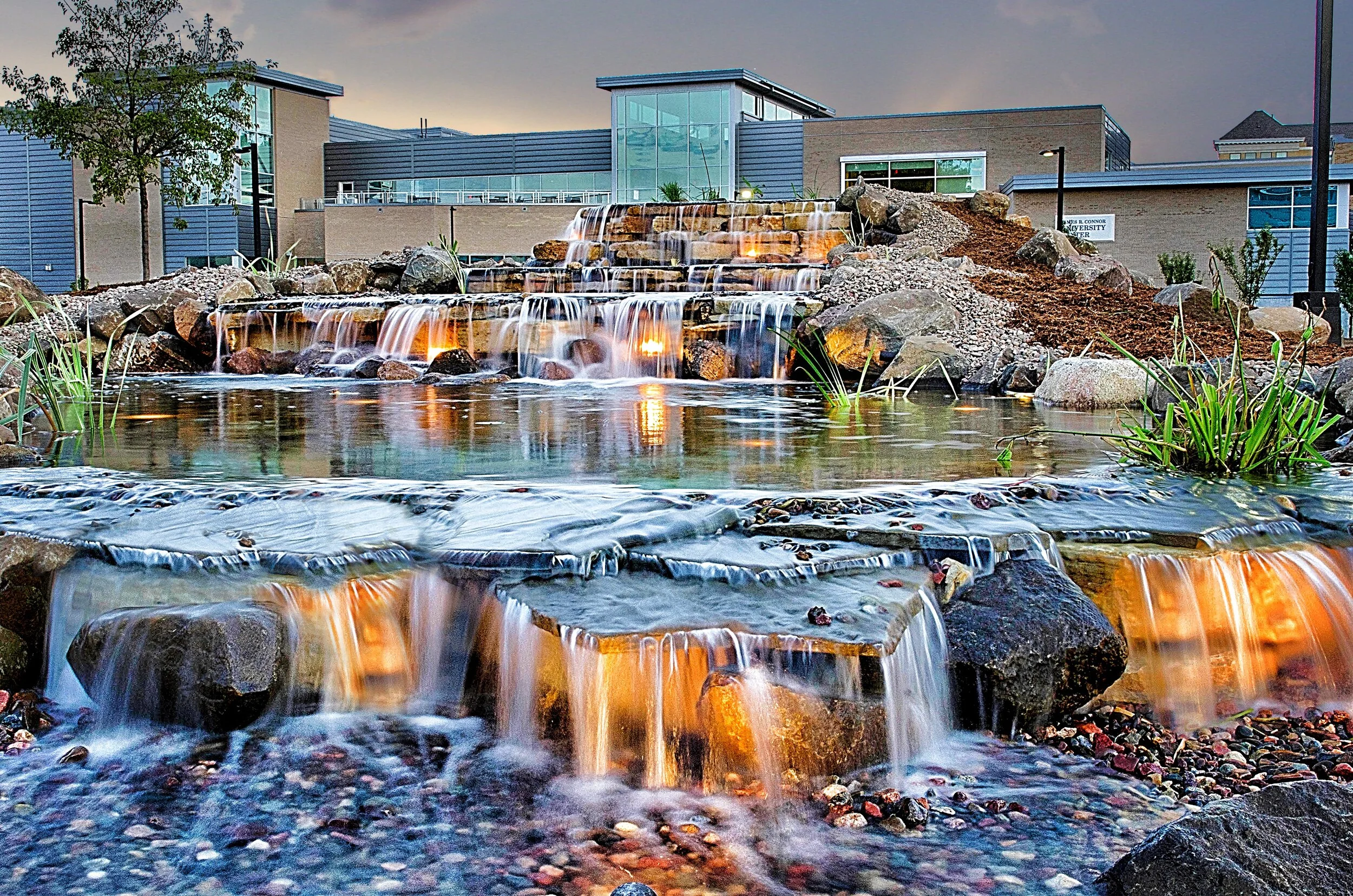 A modern building with large glass windows and an outdoor decorative water feature with small waterfalls and rocks, illuminated by underwater lighting, surrounded by plants and trees.