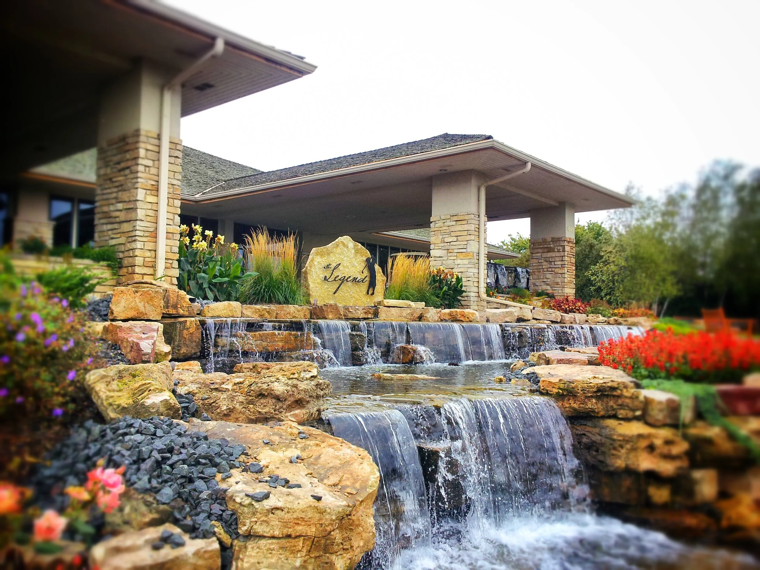 A landscaped yard featuring a water fountain cascading over rocks, surrounded by colorful flowers and greenery, with a modern house in the background.