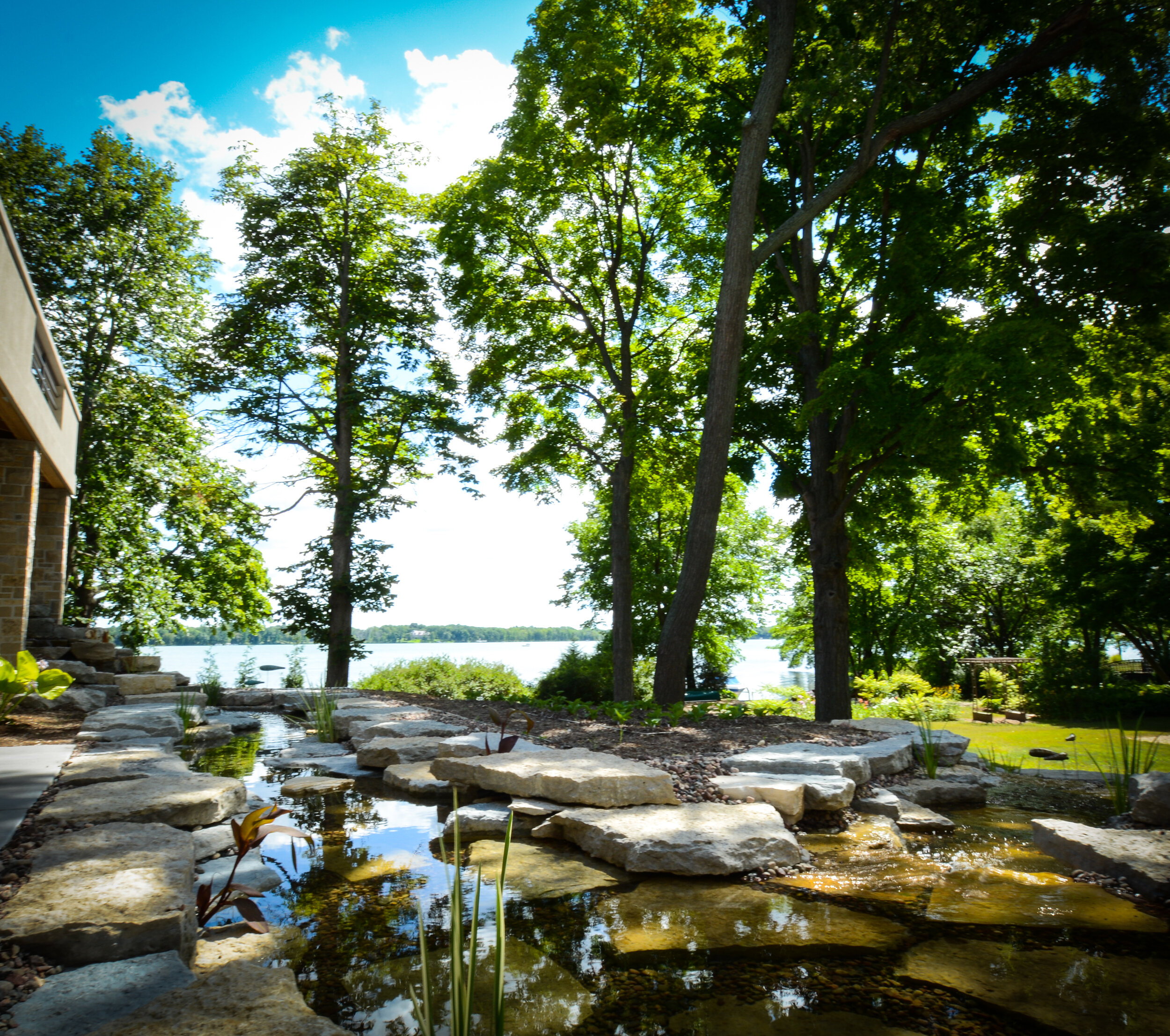 A backyard garden with a small pond edged with rocks, surrounded by trees and lush greenery, with a view of a lake and blue sky with white clouds in the background.