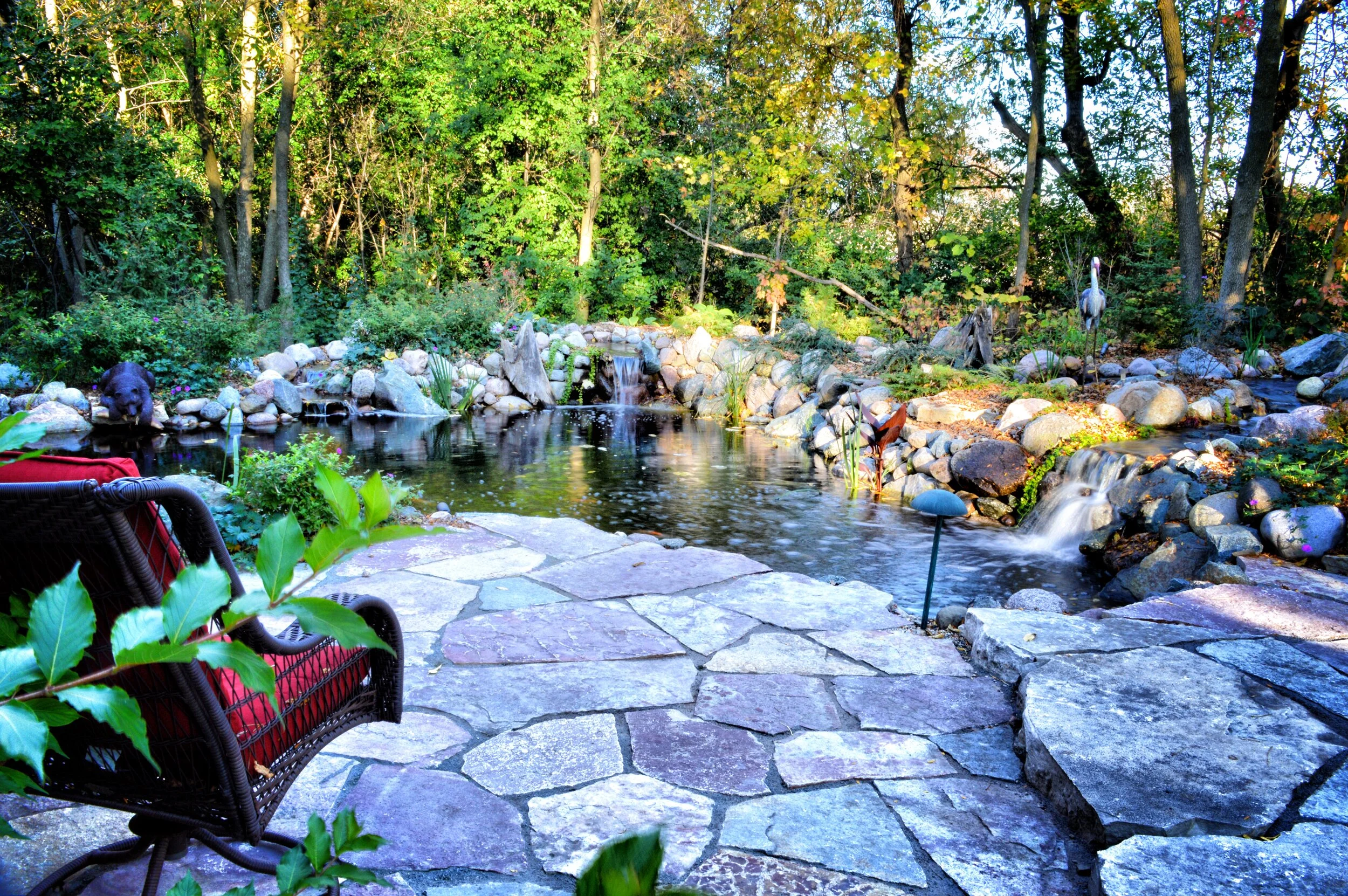 A backyard garden with a stone patio overlooking a pond surrounded by rocks, trees, and plants, with a shed or fence in the background.