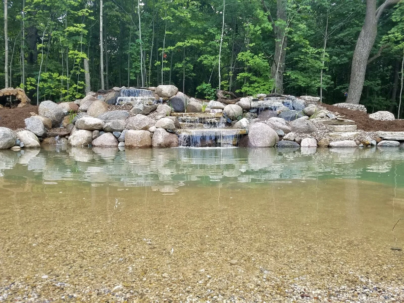 A small pond with a rocky waterfall feature in a wooded area, with it flowing down multiple levels of rocks into the pond.