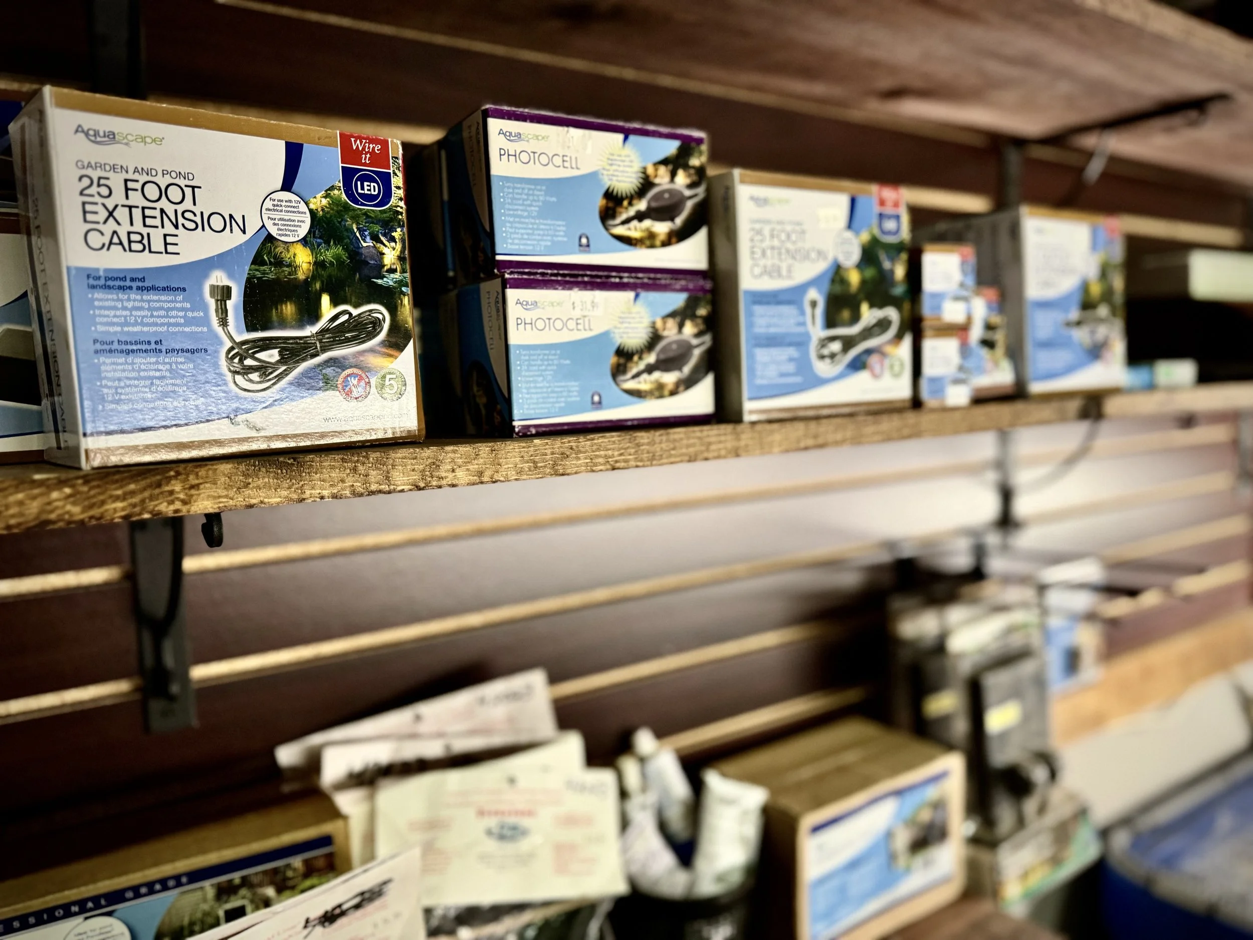 A wooden shelf stocked with boxes of gardening supplies, including extension cables and photochemical products.