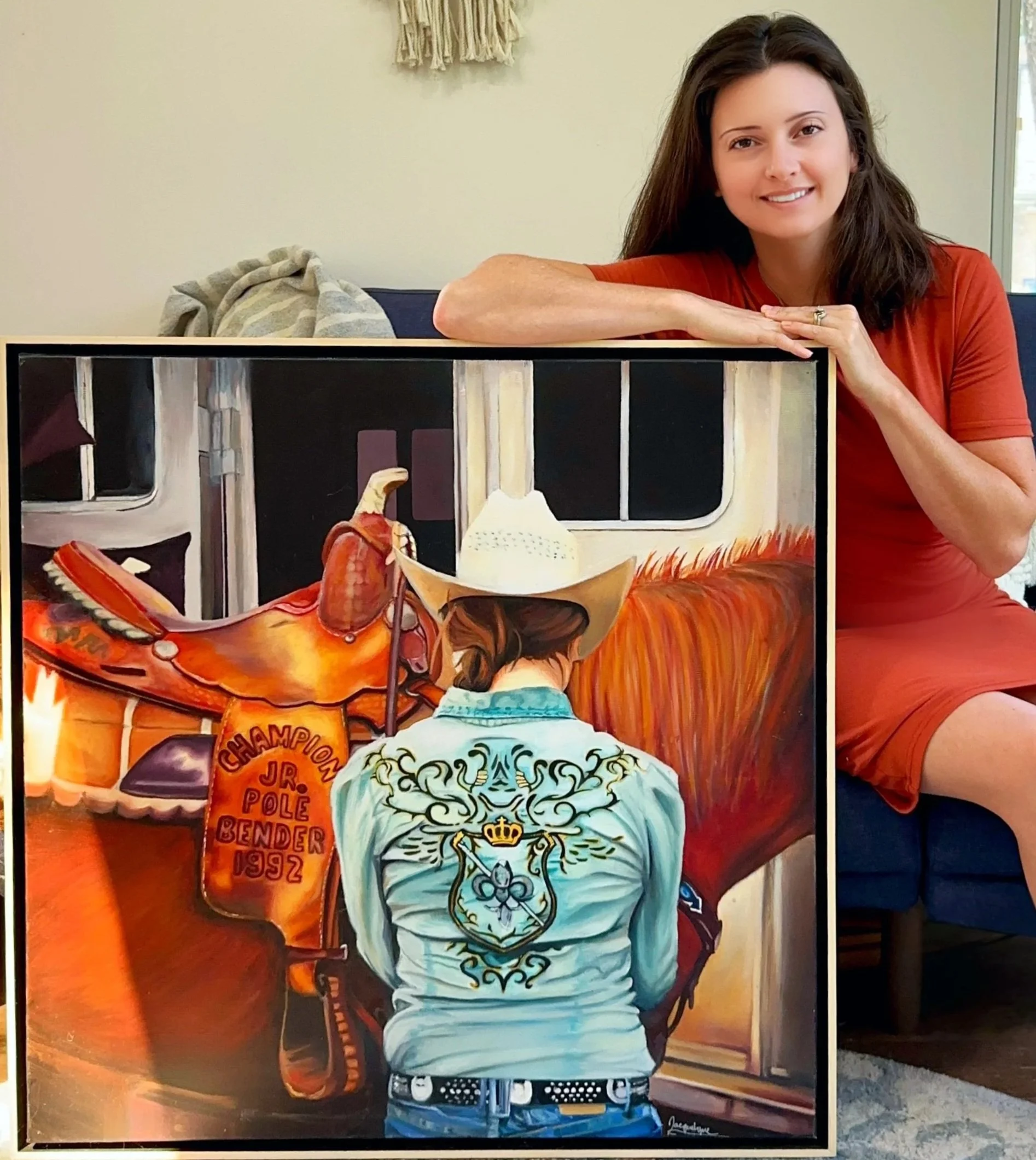 A woman in a red dress sitting on a blue sofa, smiling, with her left arm resting on a framed painting of a cowboy from behind inside a train.