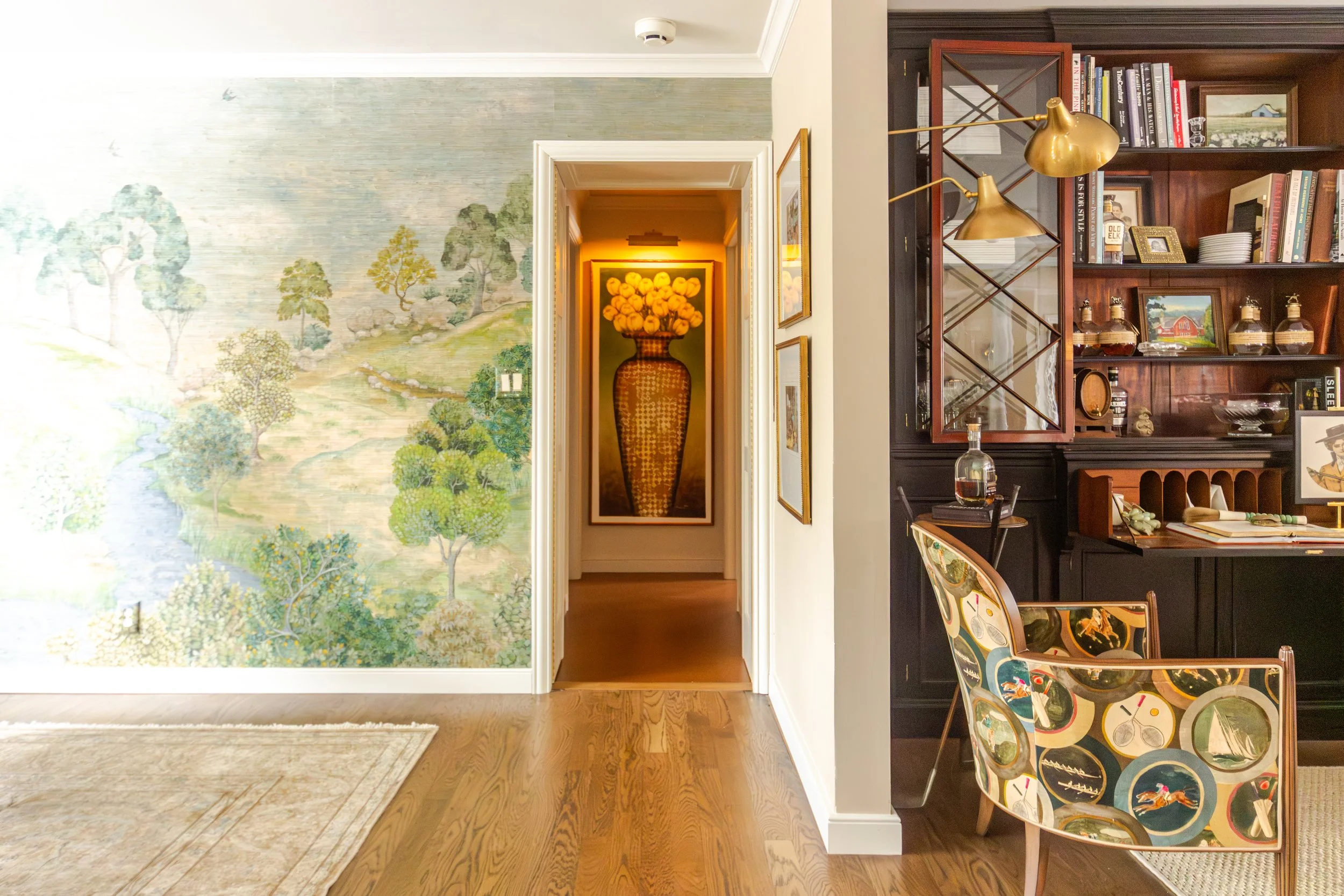 A cozy study room with a nature mural on one wall and a hallway with a flower painting visible. A vintage chair and wooden bookshelf add warmth.