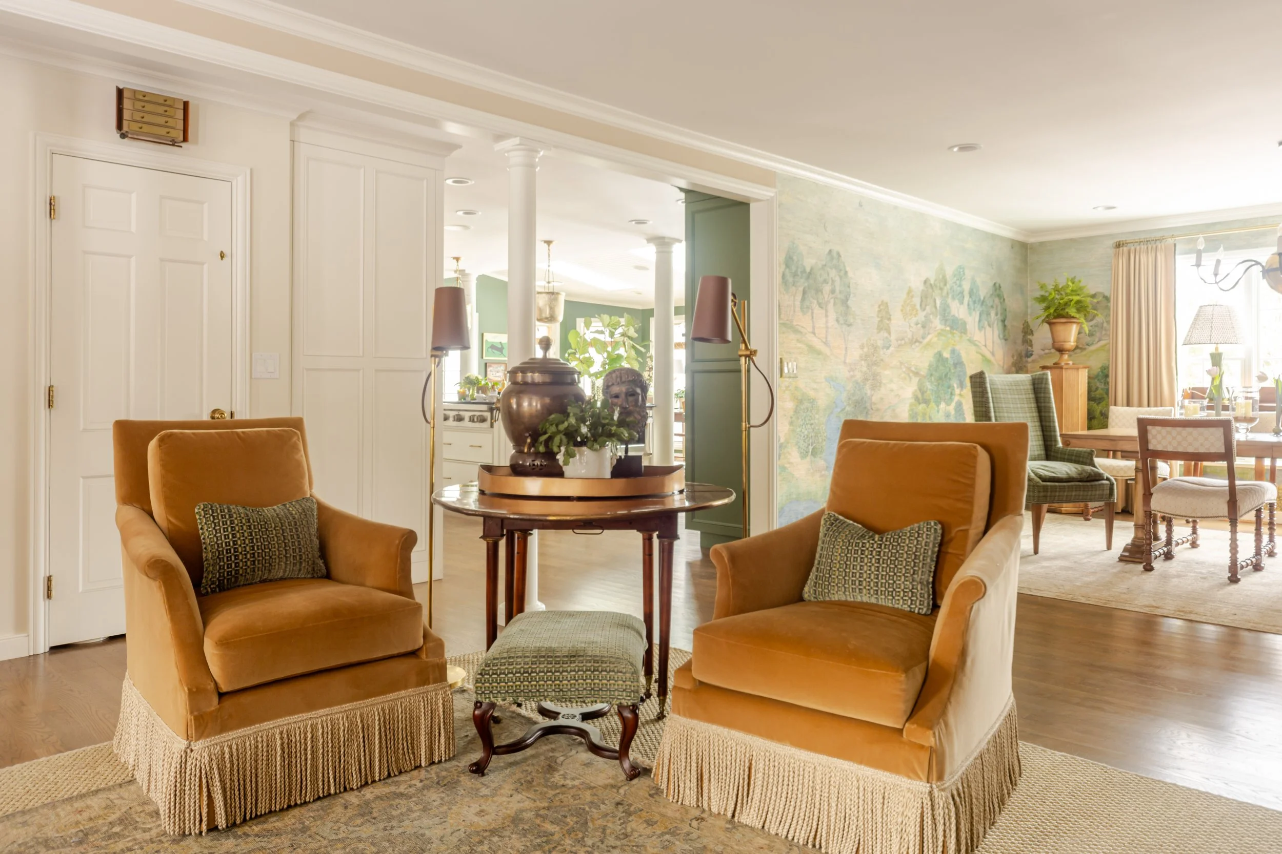 Warm, elegant living room with two mustard velvet armchairs, green cushions, round table, and lush plants. Mural wall and dining area in background. Cozy ambiance.