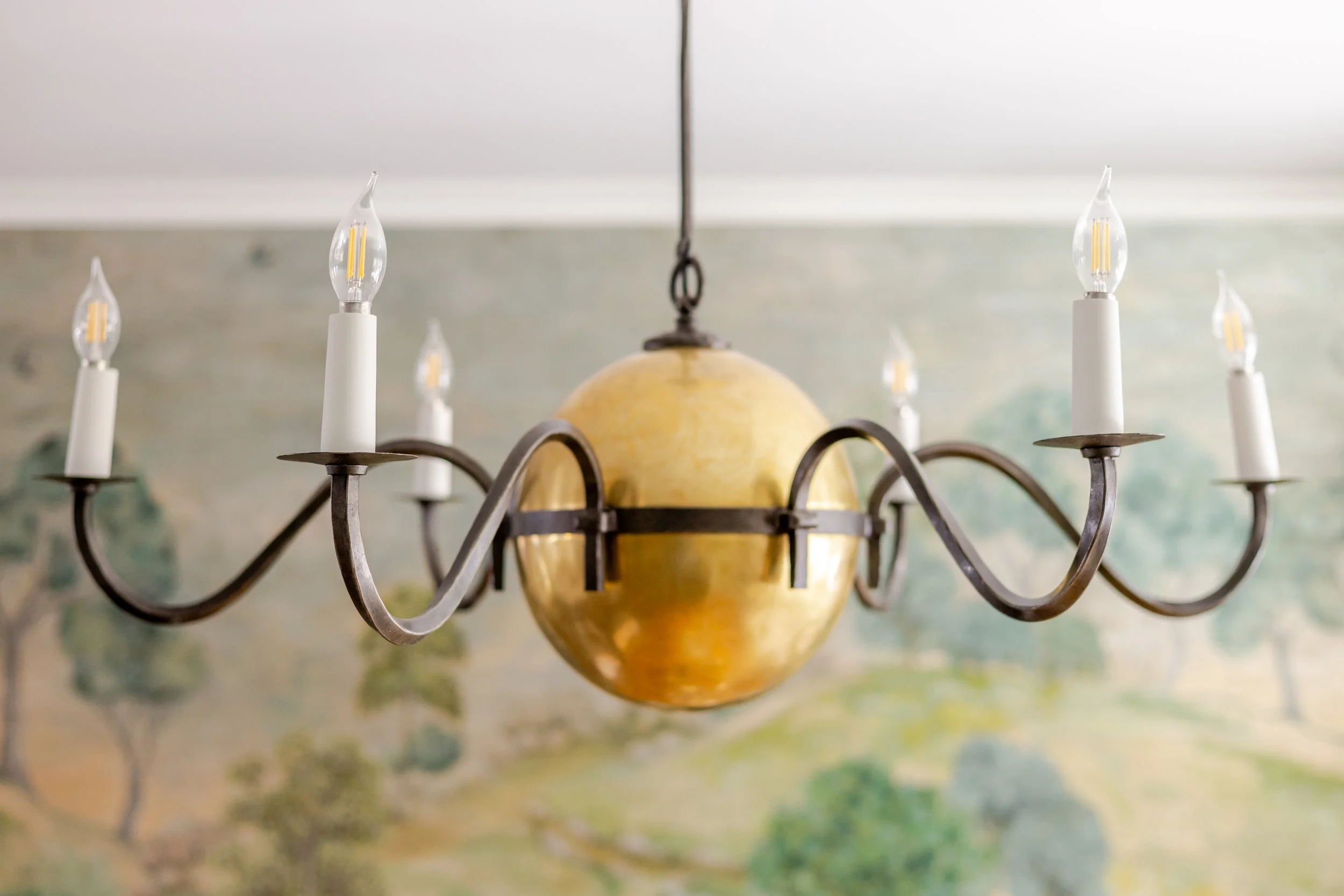Close-up of a sculptural chandelier featuring a warm brass globe center and gracefully curved dark metal arms with candle-style bulbs. The fixture hangs against a softly painted landscape mural, blending classic craftsmanship with artistic detail.