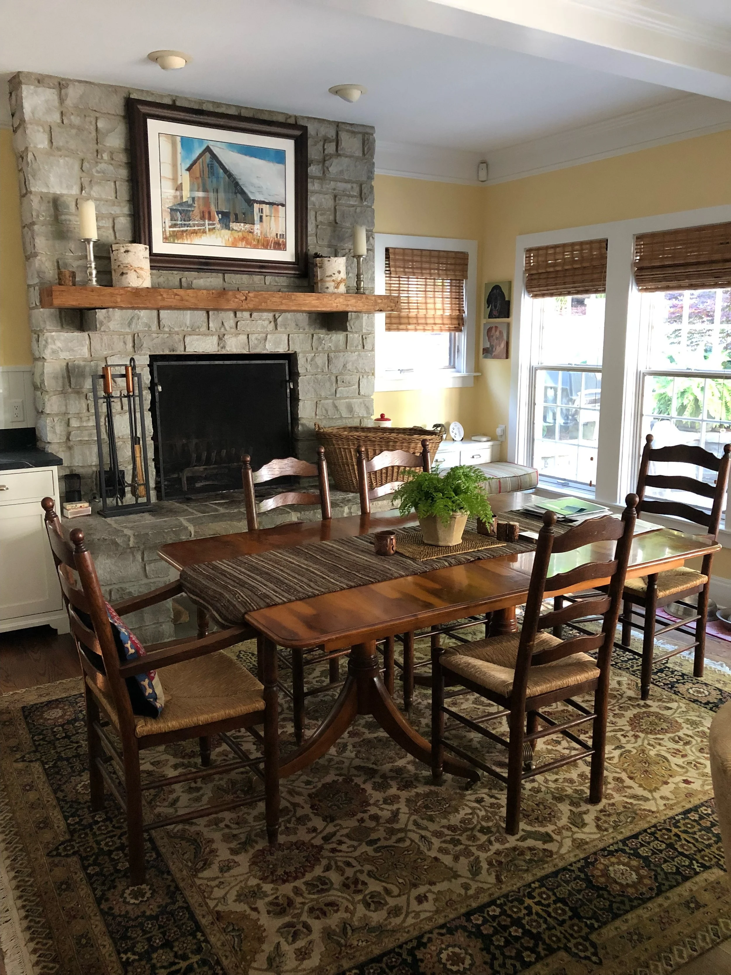Traditional dining area with a wooden table and chairs, stone fireplace, rustic mantel, woven window shades, and warm natural light.