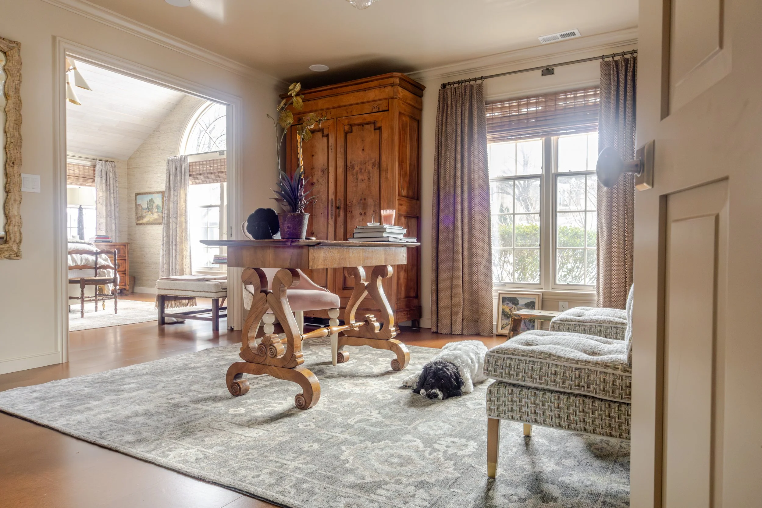 Cozy room with wooden furniture, large window draped with curtains, and patterned rug that leads into a primary bedroom. A small dog lies on the rug, creating a peaceful, warm atmosphere.