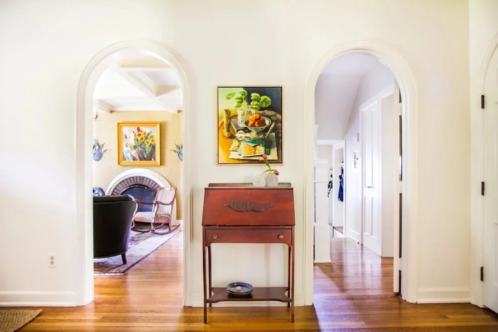 Bright hallway with arched doorways, antique wooden secretary desk, framed still-life artwork, and warm hardwood floors connecting classic living spaces.