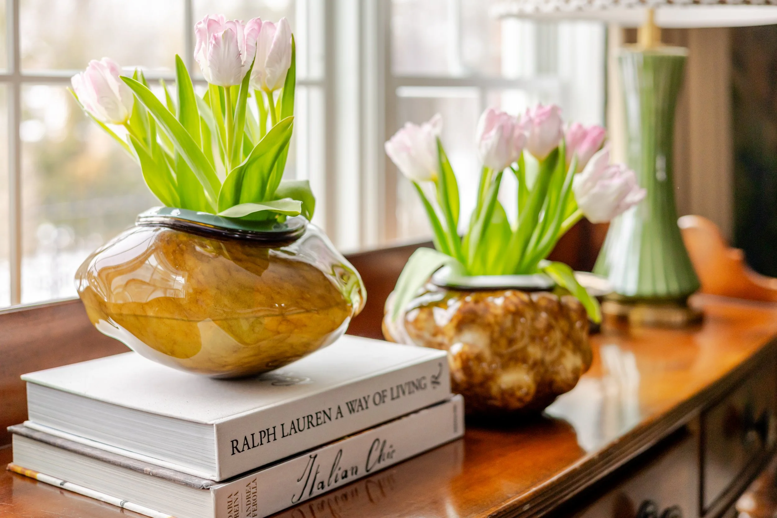 Styled vignette featuring pale pink tulips arranged in sculptural amber glass vessels atop stacked design books on a polished wood console. Soft natural light from nearby windows highlights the organic textures and refined, curated aesthetic.