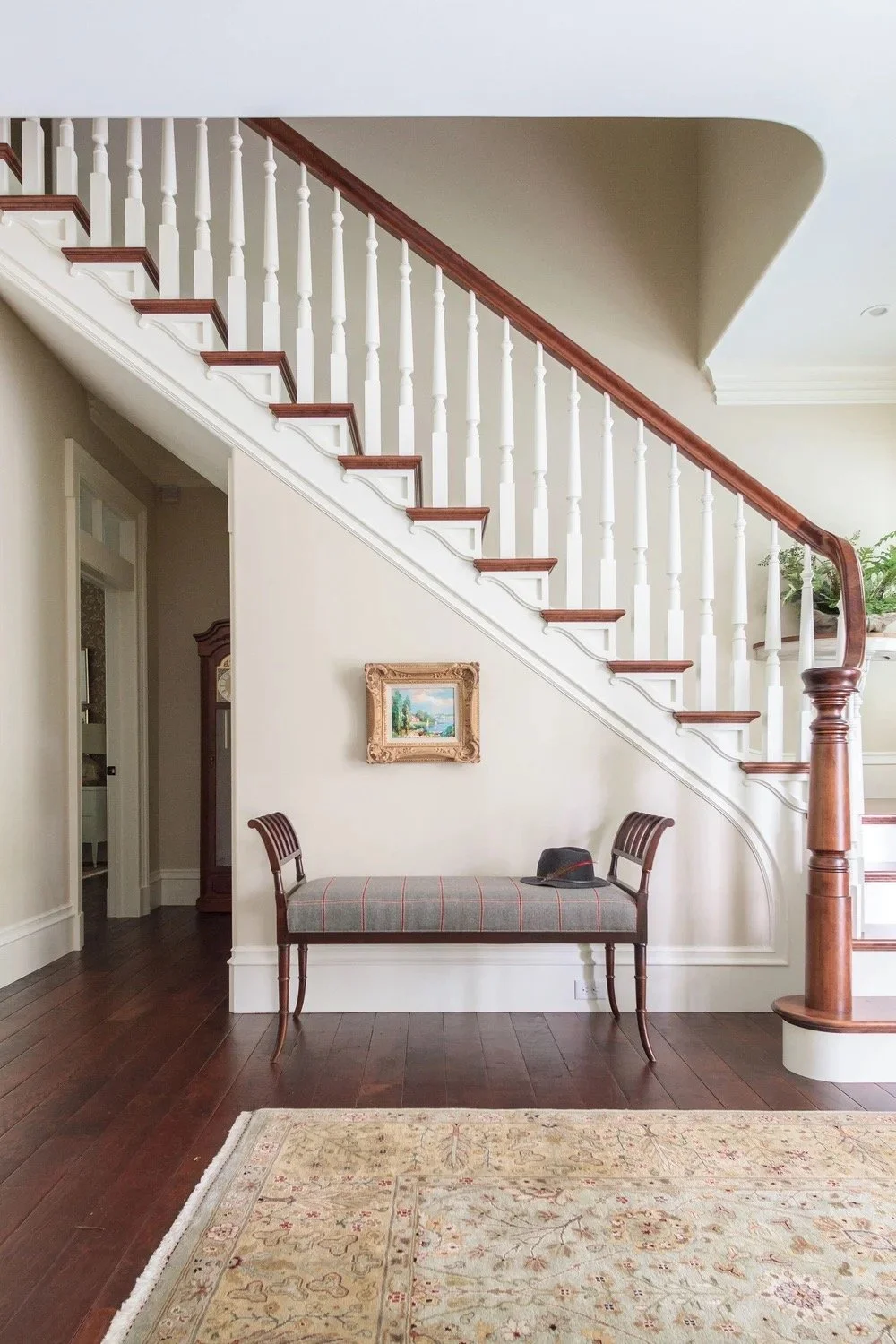 Elegant foyer with a round pedestal table, large indoor plant, wooden staircase, bench seating, and natural light over hardwood floors.