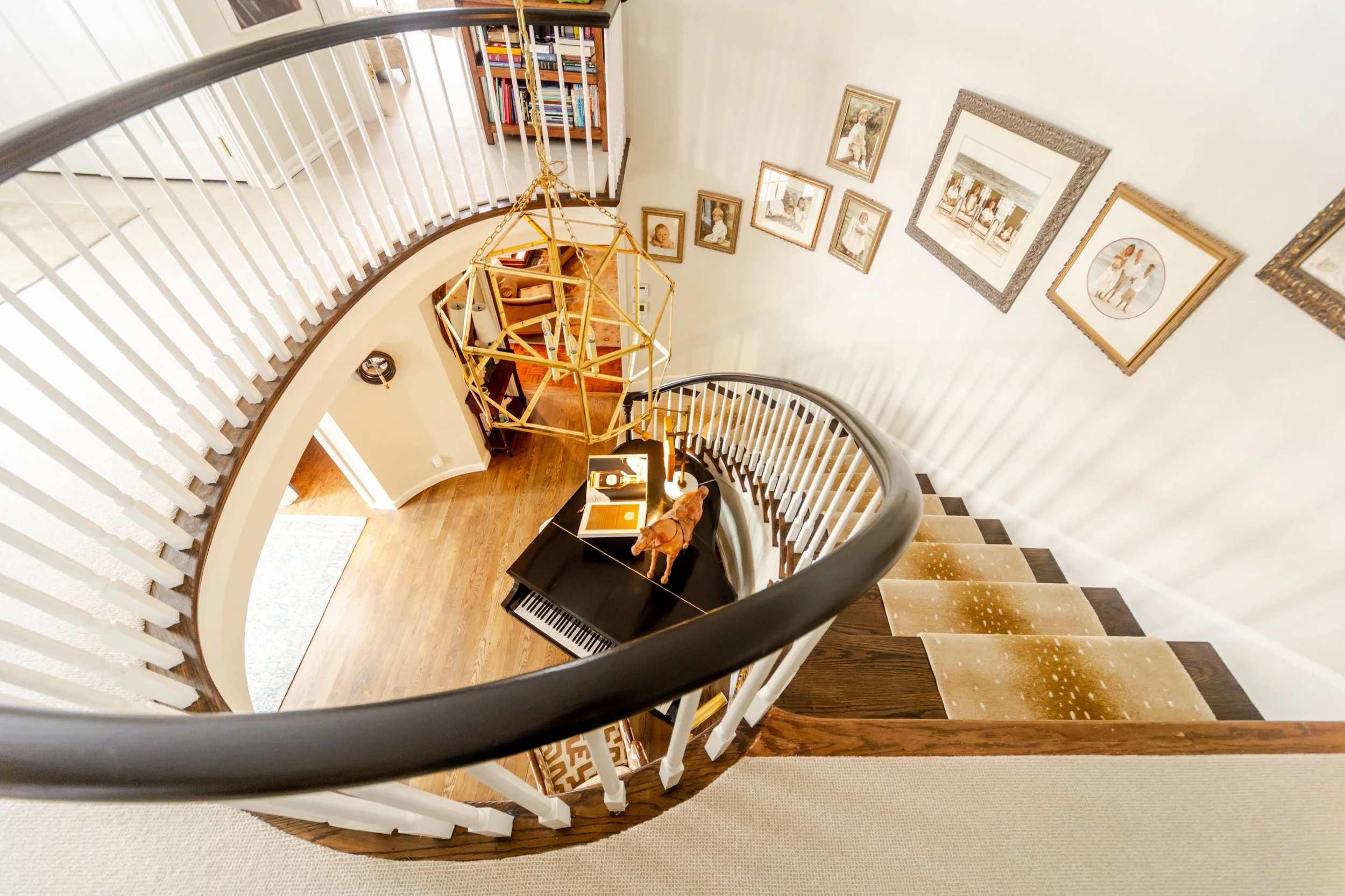 A spiral staircase with dark wood railing curves around a geometric chandelier. Framed art decorates the wall, and a piano sits on the wooden floor below.