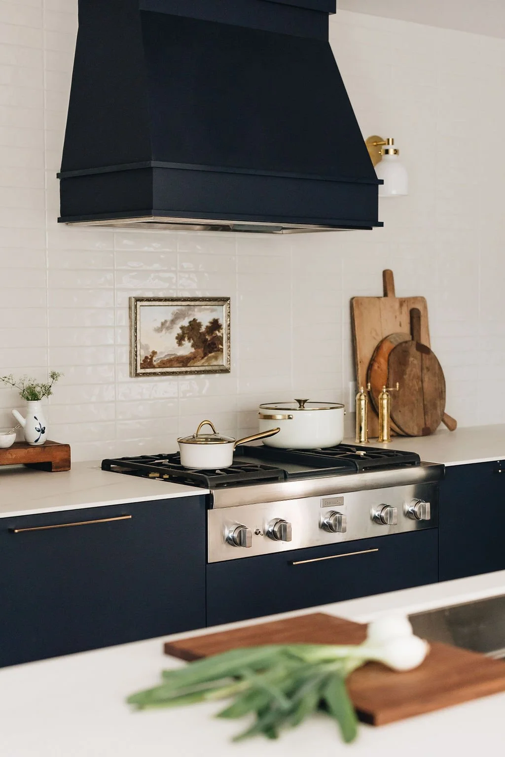 Historic home kitchen renovation by Mimi Pineau Design in Toronto features a gas range top and vent hood nestled into dark blue cabinetry; cream tile backsplash, white sconces, brass hardware and rustic accessories complement the decor.