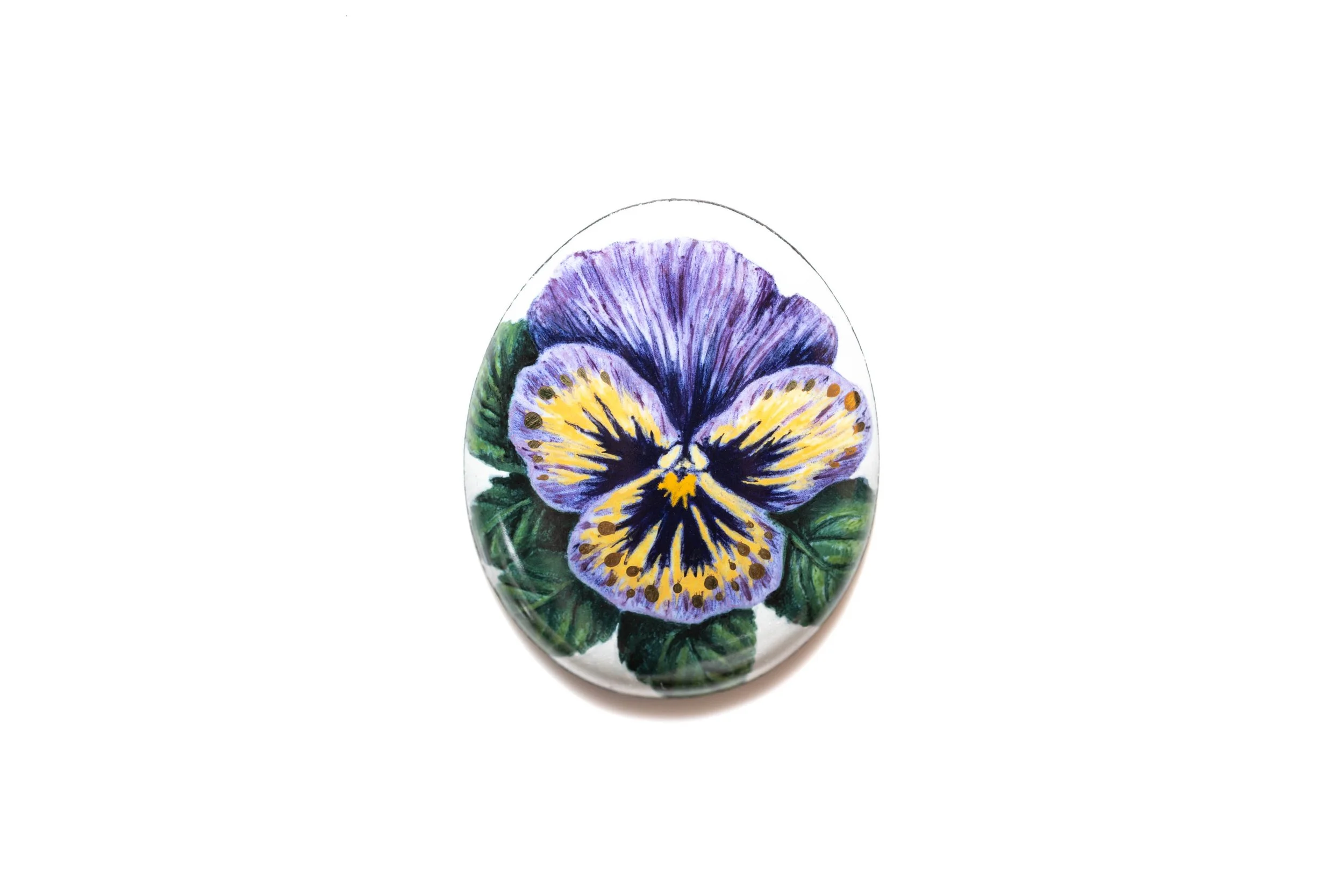 Purple and yellow pansy with green leaves, hand-painted in vitreous enamel on an oval cabochon.