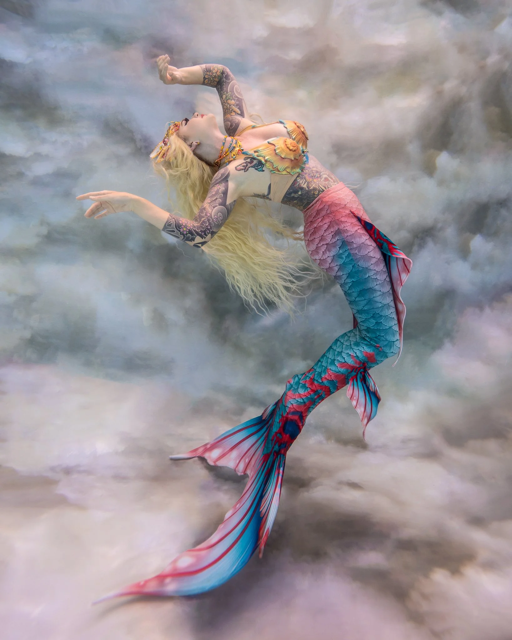 Professional mermaid Haley poses underwater in a heated pool in Orlando, wearing a beautiful pink and blue tail, and some shells, with long flowing blonde hair.