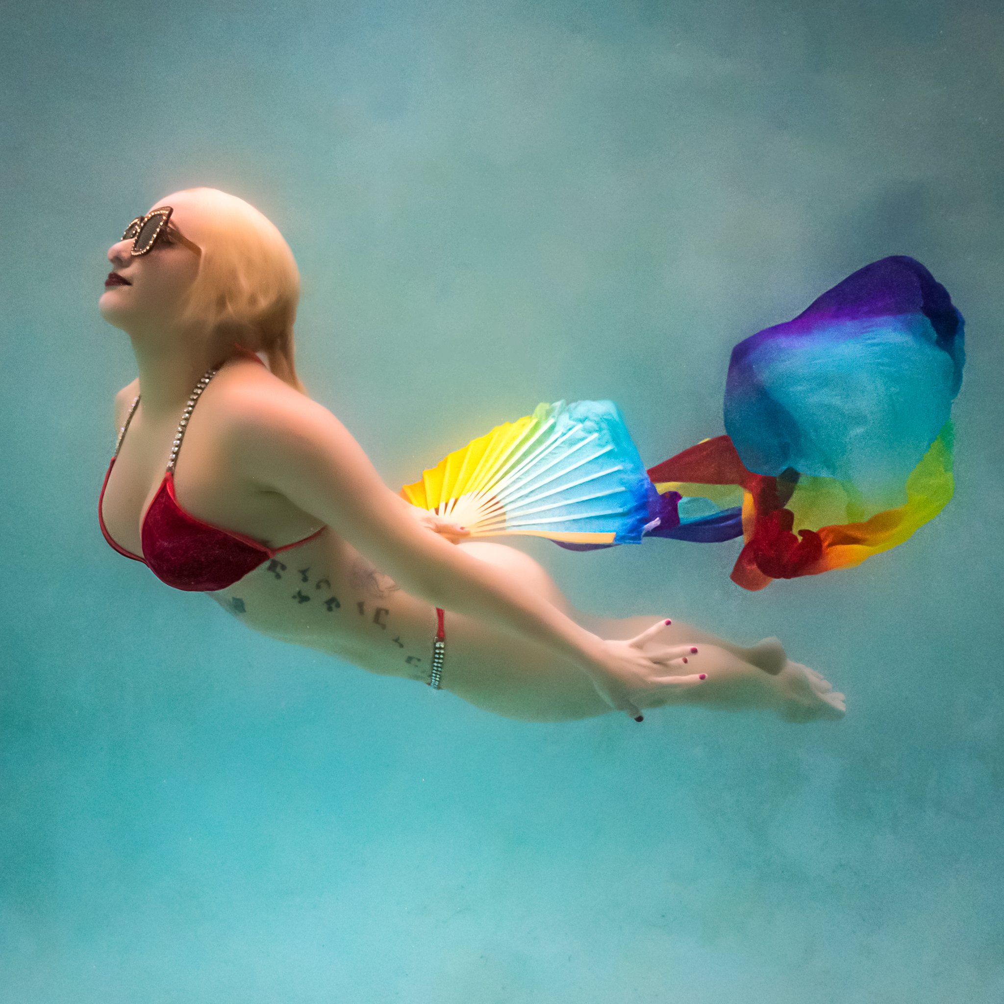Amanda swims in a red bikini and sunglasses with a rainbow fan underwater in a heated pool in Orlando, FL
