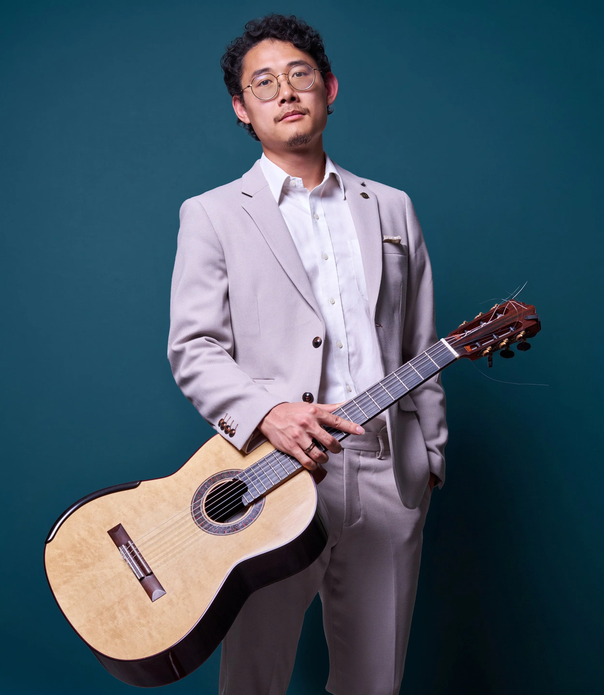 A man in glasses and a light gray suit holding a guitar against a dark teal background.