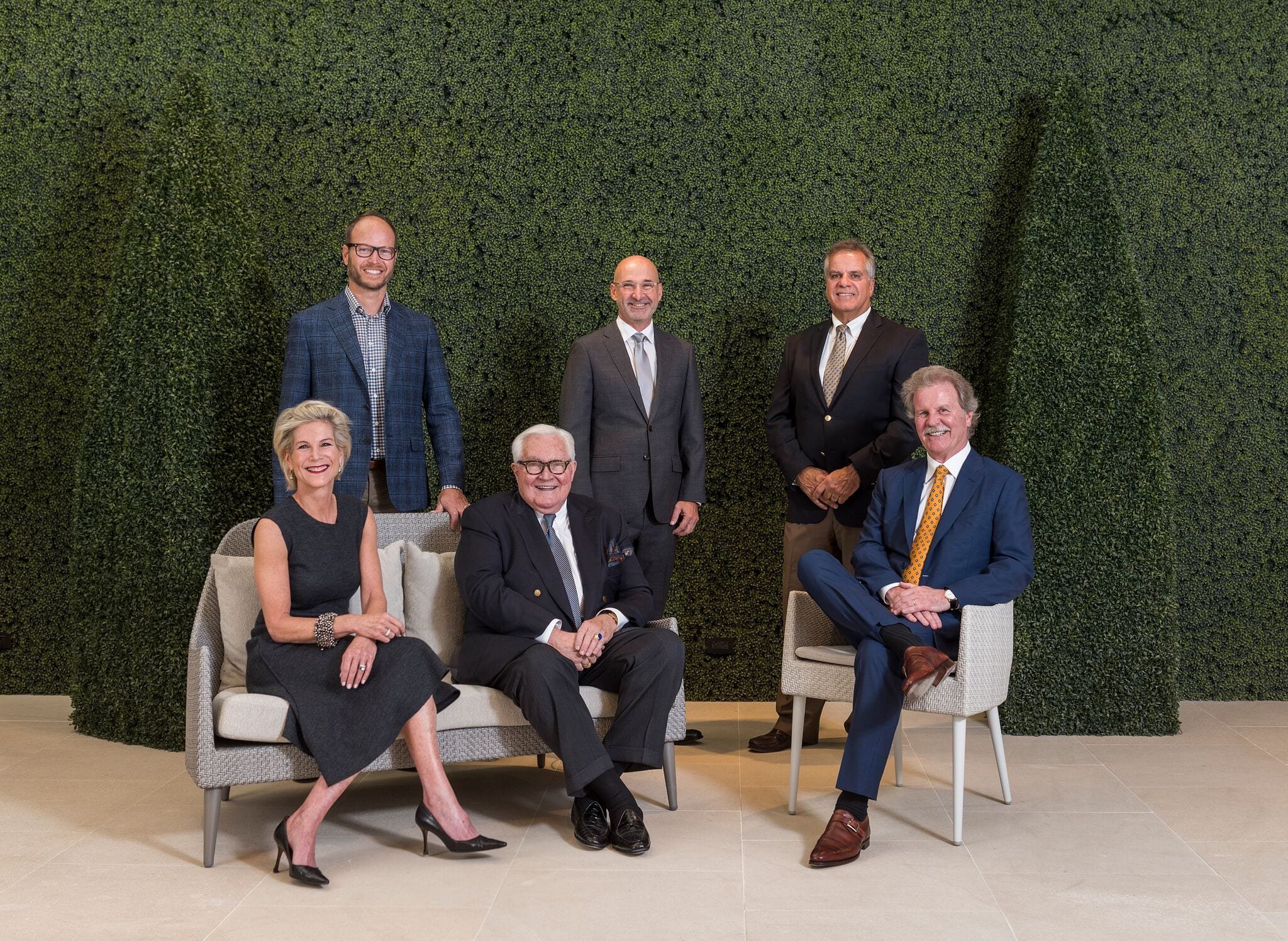 Photo of six people, five men and one woman dressed in professional attire posing in front of a green foliage backdrop.