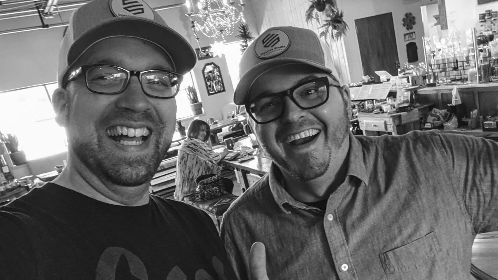 Two men wearing glasses and baseball caps smiling at the camera in a restaurant or café. A woman is sitting at a table in the background, and the decor includes Christmas ornaments.