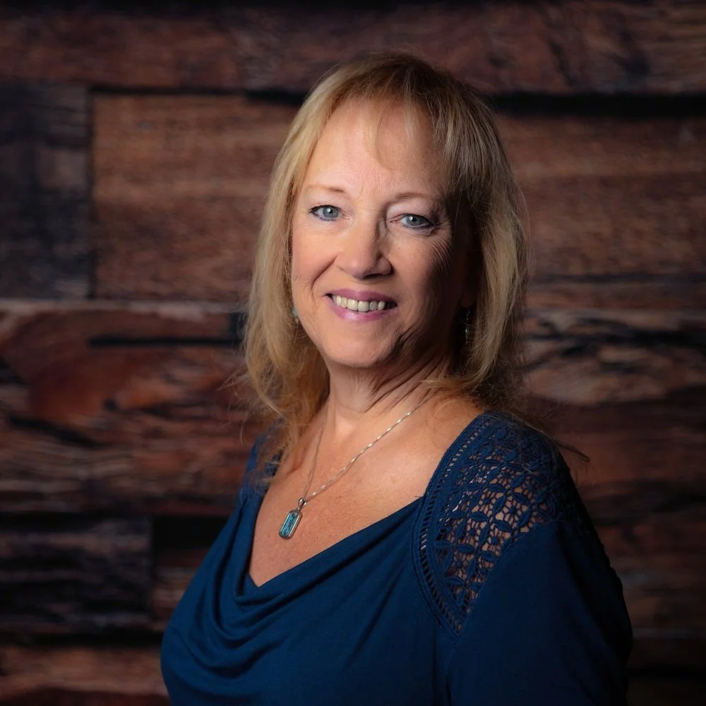 A smiling middle-aged woman with blonde hair and blue eyes, wearing a blue top with lace details on the shoulders, standing in front of a wooden background.