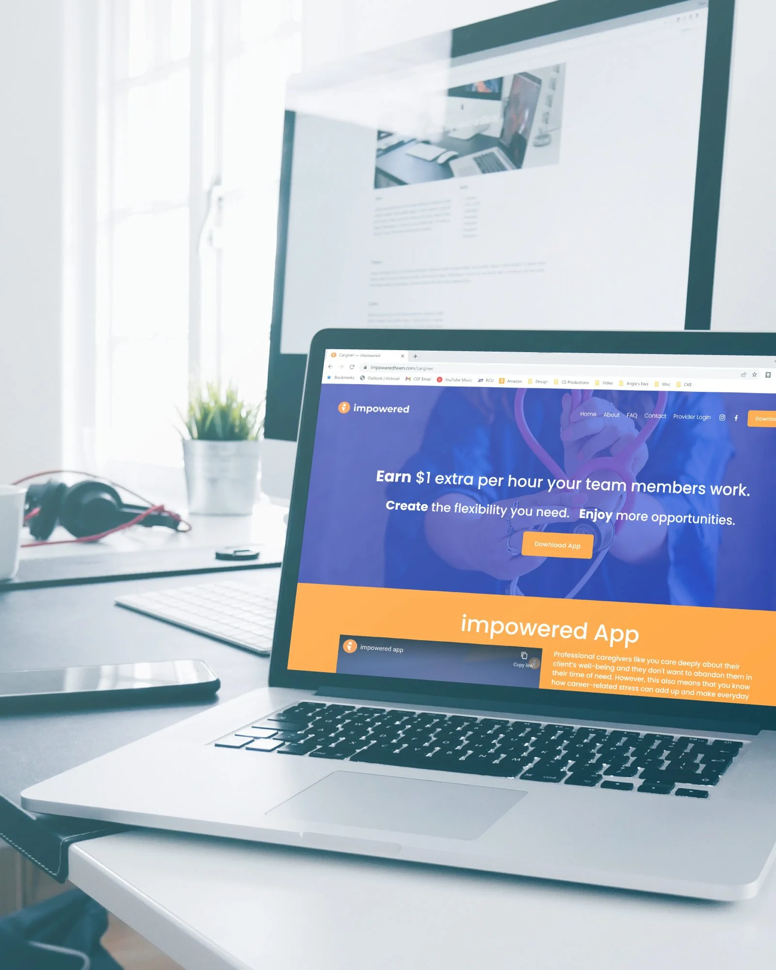 A laptop on a white desk displaying the Empowered app website, with a larger monitor in the background, a plant, headphones, a smartphone, and a keyboard nearby.