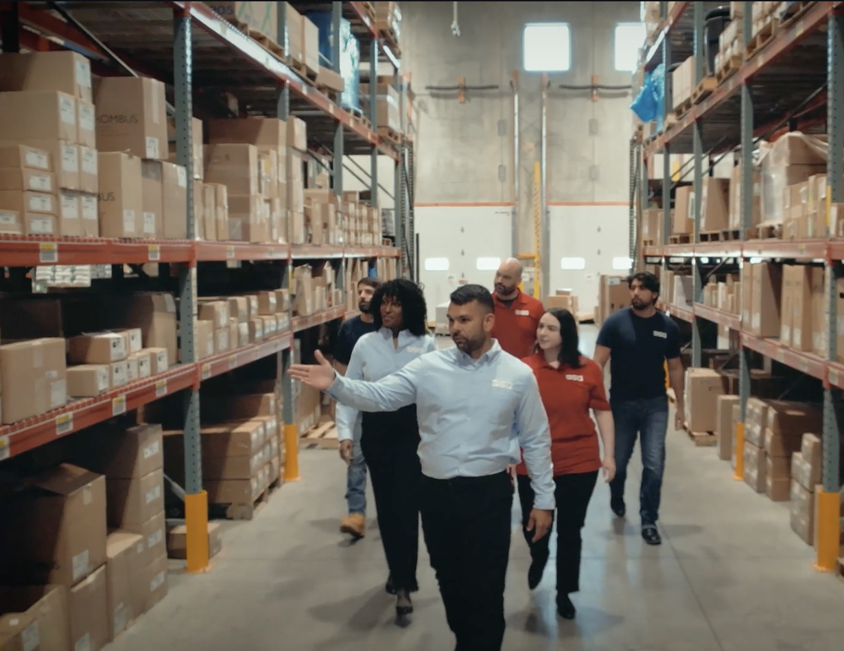 Group of six people walking through a warehouse aisle with shelves filled with boxes.