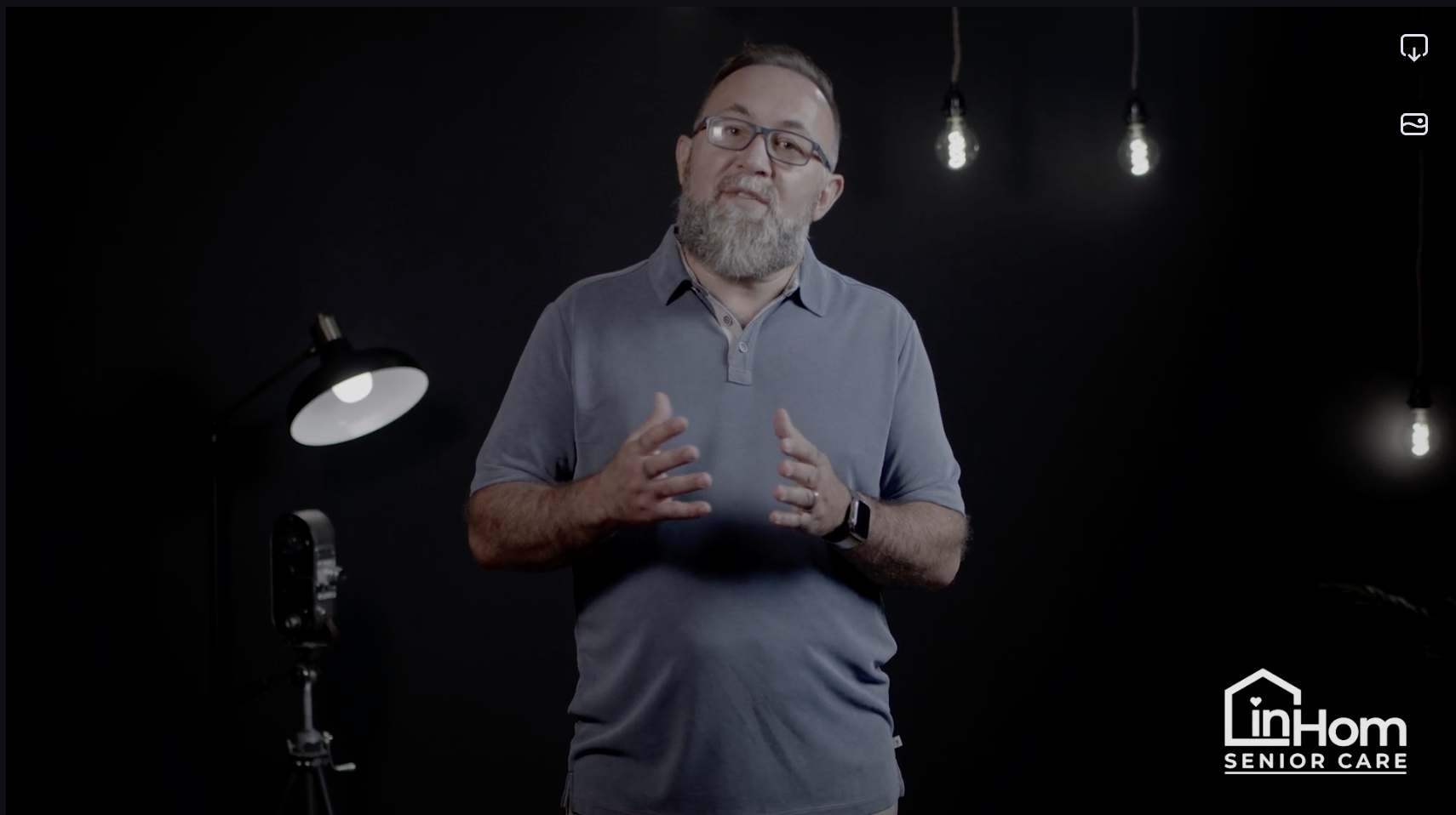 A man with glasses, gray beard, wearing a gray polo shirt, standing in front of a dark background, speaking with lighting equipment and hanging light bulbs behind him. A logo in the bottom right corner reads "inHom Senior Care."